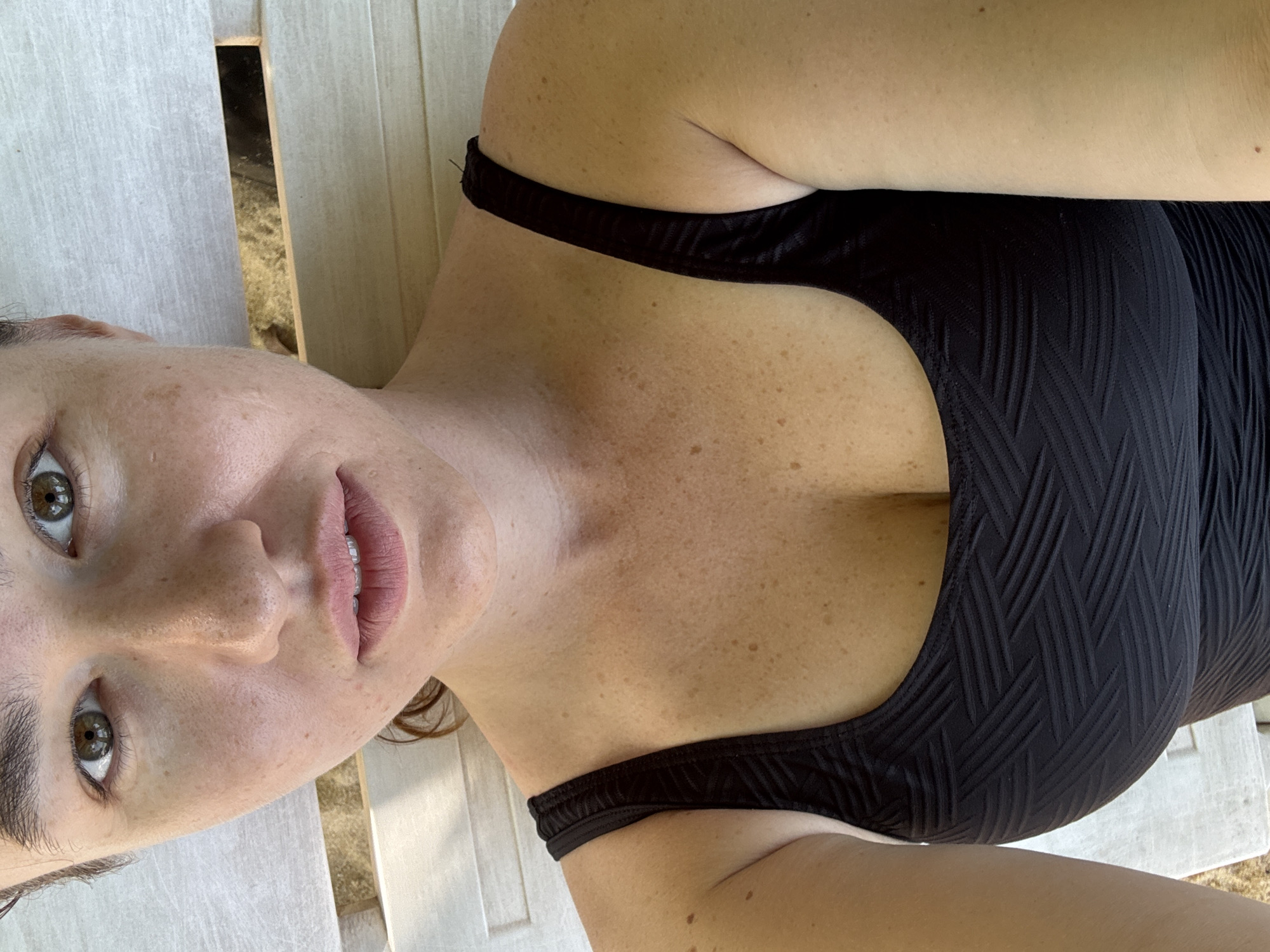 In Rawai, Thailand, a close-up, overhead shot shows a woman wearing a black textured swimsuit. Her eyes are green, and she has a fair complexion with freckles across her face and chest. Her lips are pink and slightly parted, revealing her teeth. The background consists of light-colored wooden planks, suggesting an outdoor setting.