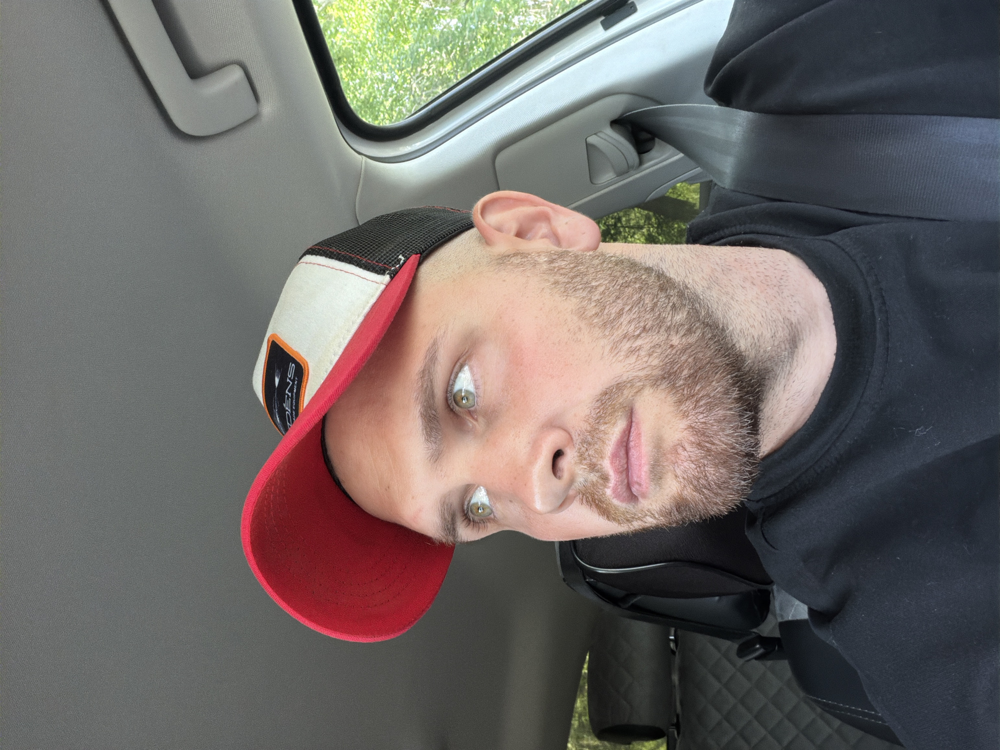 The image displays a male individual from the chest up, positioned inside what appears to be a vehicle. His head is tilted to the right, and he is looking directly into the camera. He has light-colored eyes (appearing hazel or green) and a short, light brown beard. He is wearing a black hoodie or sweatshirt and a baseball cap with a red brim, a white front panel, and a black mesh back. A black seatbelt is visible across his chest.
The interior of the vehicle features gray paneling on the ceiling and side, along with a gray grab handle. A window reveals green foliage, suggesting an outdoor, daytime setting. The seat behind the subject has a dark, quilted pattern. A patch on his cap clearly displays the text "ODENS" on the top line and "GEAR EQUIPMENT" on the bottom line. The overall image is rotated 90 degrees counter-clockwise.