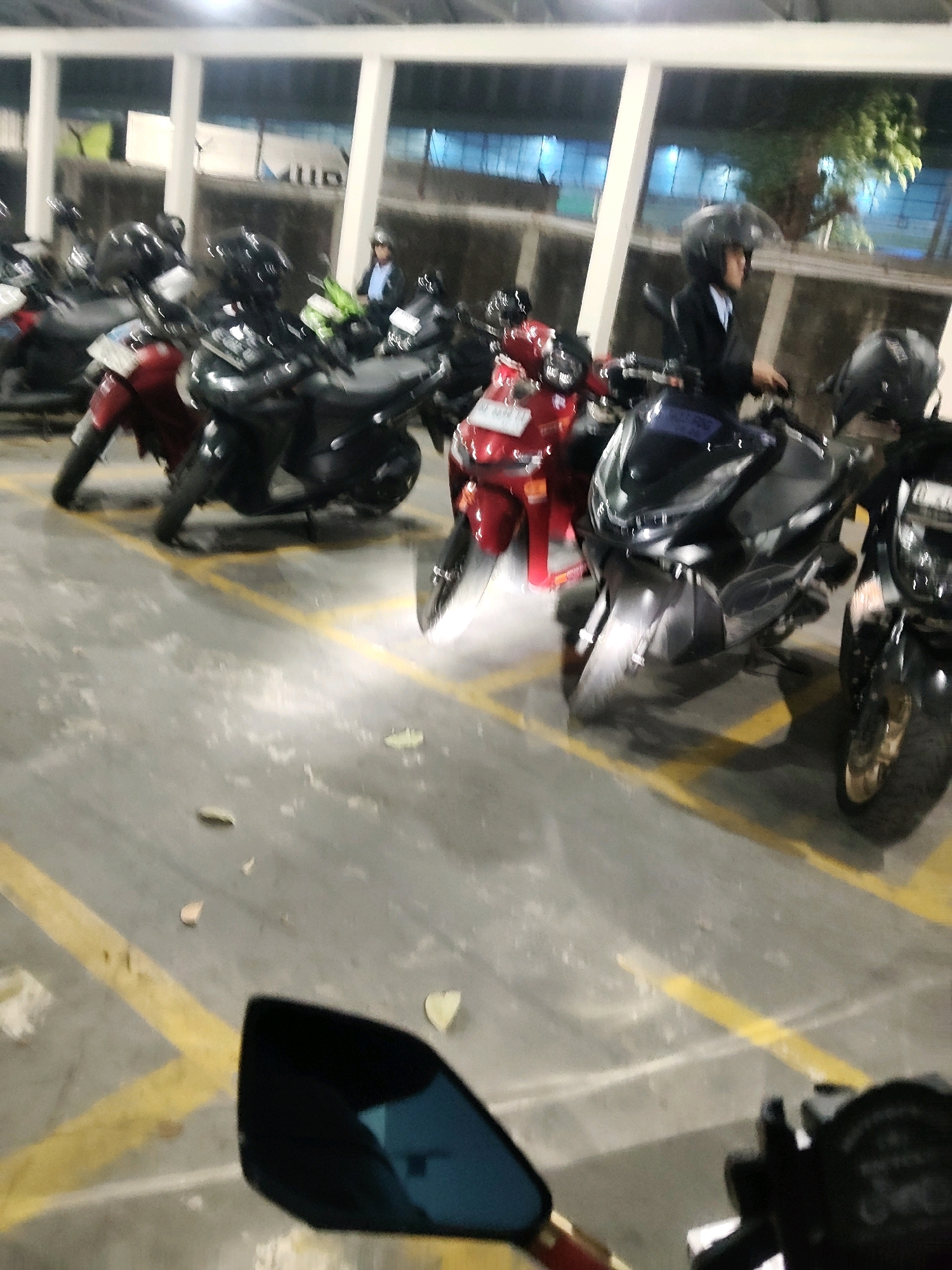 A person wearing a black helmet and a blue collared shirt with a black jacket is seated on a black motorcycle in an outdoor parking lot, possibly at night or in a dimly lit area. The motorcycle is parked next to other motorcycles, including a red one and a dark gray one. The ground is paved and marked with yellow lines, suggesting designated parking spots. The background features a building structure with white vertical pillars and large windows or openings, through which some greenery and blue lighting are visible. The image is slightly blurred, indicating motion or low light conditions.