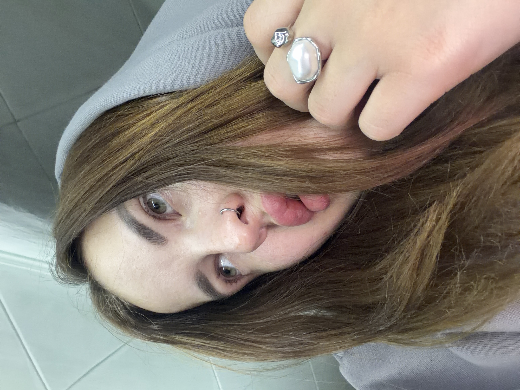 An overhead shot depicts a person with light brown hair, wearing a light gray hooded sweatshirt. The person has a silver ring with a large pearl-like stone on their finger. They have a silver nose ring. The person is sticking out their tongue slightly. The background is a light gray or white surface, possibly a tiled wall or floor. The location in the city of Port-Katon, Russia, cannot be determined from the image.