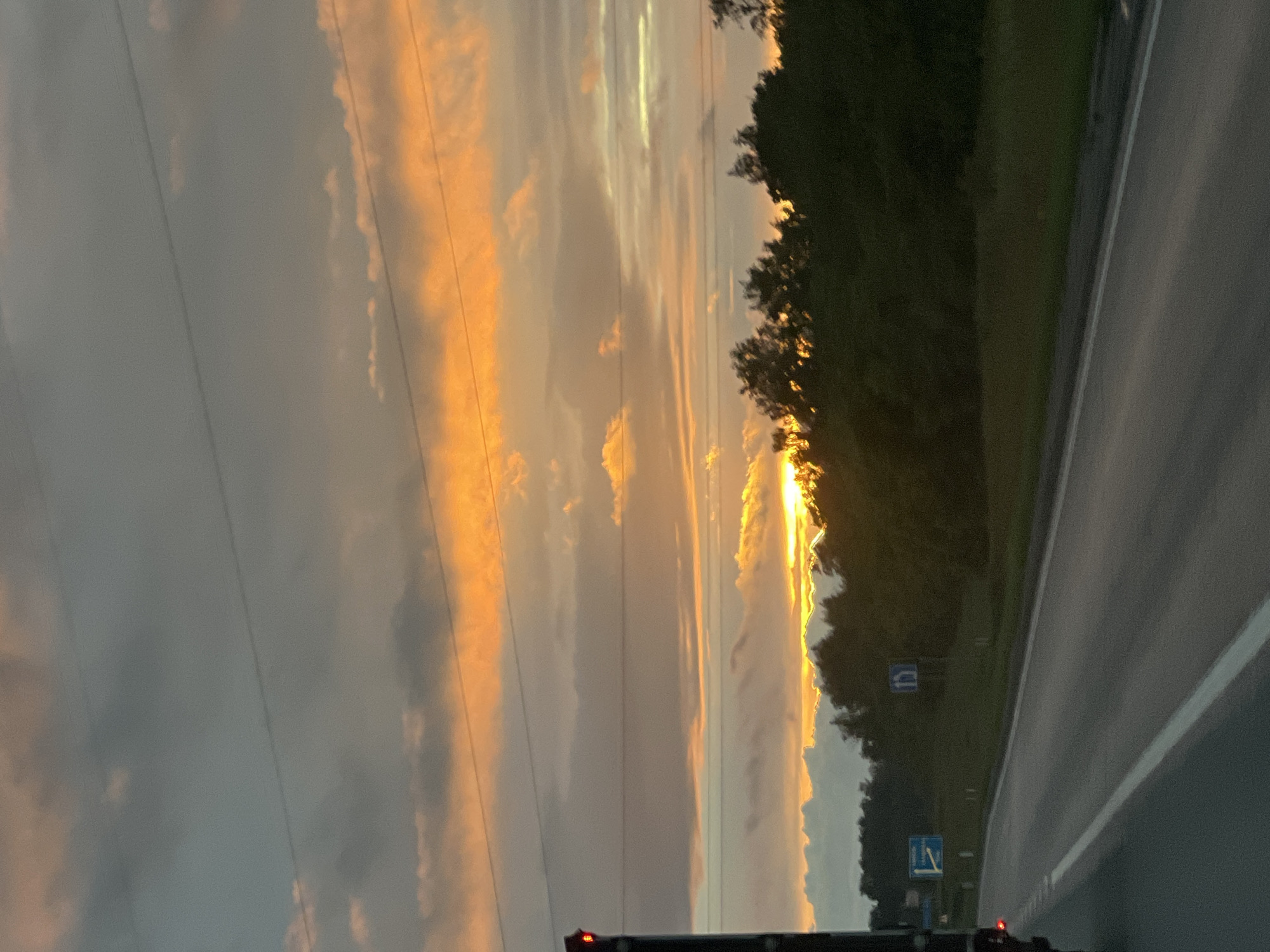 The image captures a sunset scene on a road in Kamenka, Russia. A dark-colored truck is partially visible on the left side of the frame, with its red taillight illuminated. The road stretches forward, marked with a white line. On the right side of the road, there's a line of dark green trees. The sky is filled with dramatic clouds, ranging from dark gray to vibrant orange and yellow, indicating the setting sun. Two blue road signs are visible on the roadside. One sign on the left has a white symbol and text. The other, further down on the right, displays a white arrow within a blue circle. Power lines are stretched horizontally across the upper part of the sky. The overall impression is that of a scenic drive during sunset.