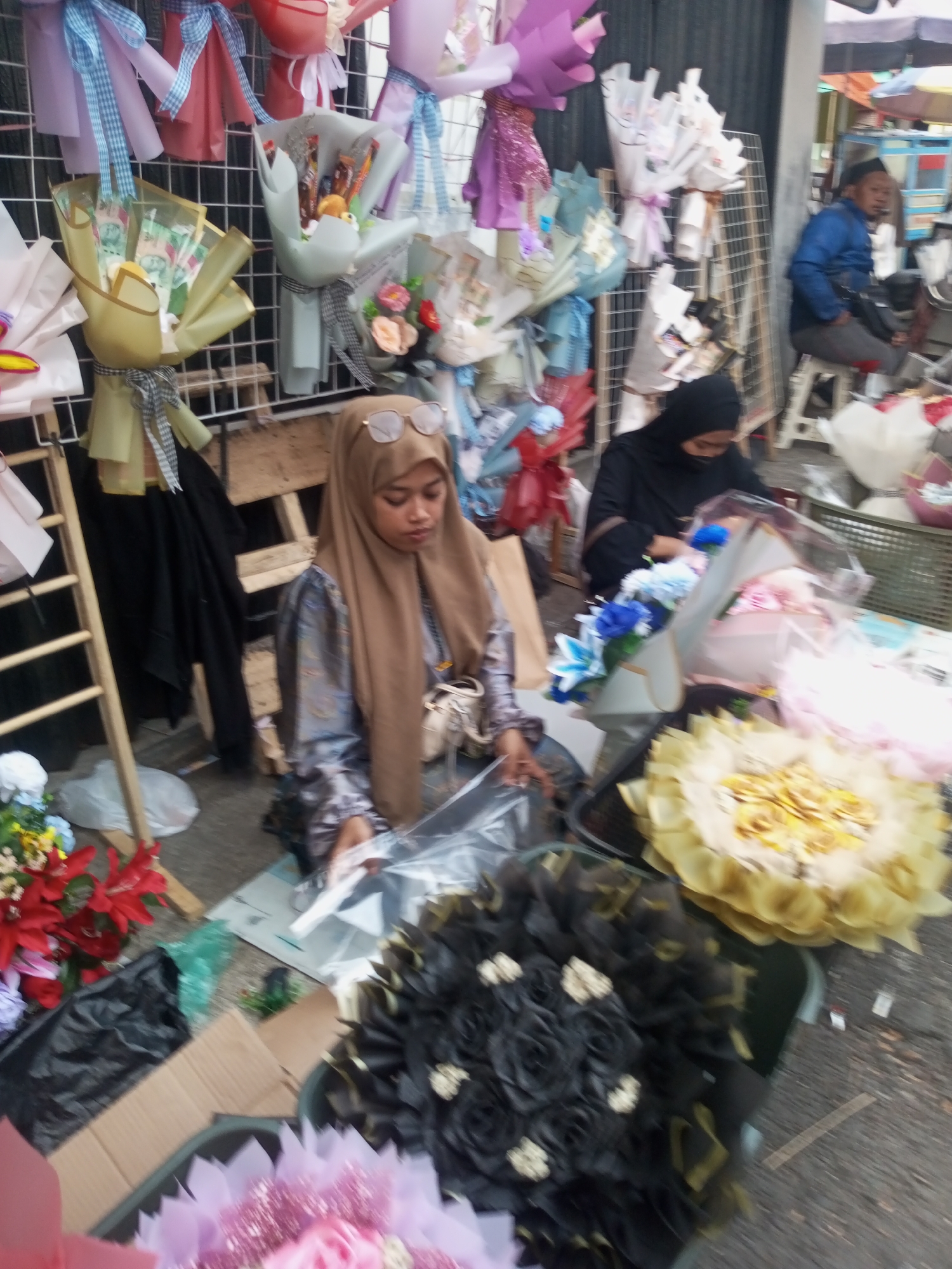 This is a medium shot, outdoor, low-angle photograph of a market stall in Pandansari, Indonesia, displaying various floral arrangements. In the foreground, a young woman wearing a hijab and glasses is seated behind a table, appearing to be arranging items. To her left are stacks of colorful artificial flowers and a ladder-like wooden structure. To her right, a man in a blue jacket and dark pants sits on a stool, facing away from the camera, while further back another person in a black hijab is visible. The stall is laden with bouquets of flowers, many of which are artificial and wrapped in colorful paper. Some bouquets contain what appear to be money and candy. The ground surface is paved. The overall scene suggests a bustling market environment.