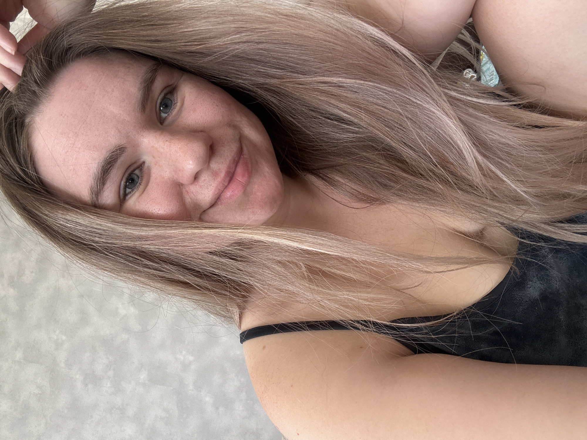 A selfie of a young woman with long, light brown hair. She is smiling slightly, looking at the camera. She is wearing a black tank top. The background is a gray, textured surface. The location cannot be determined from the image.