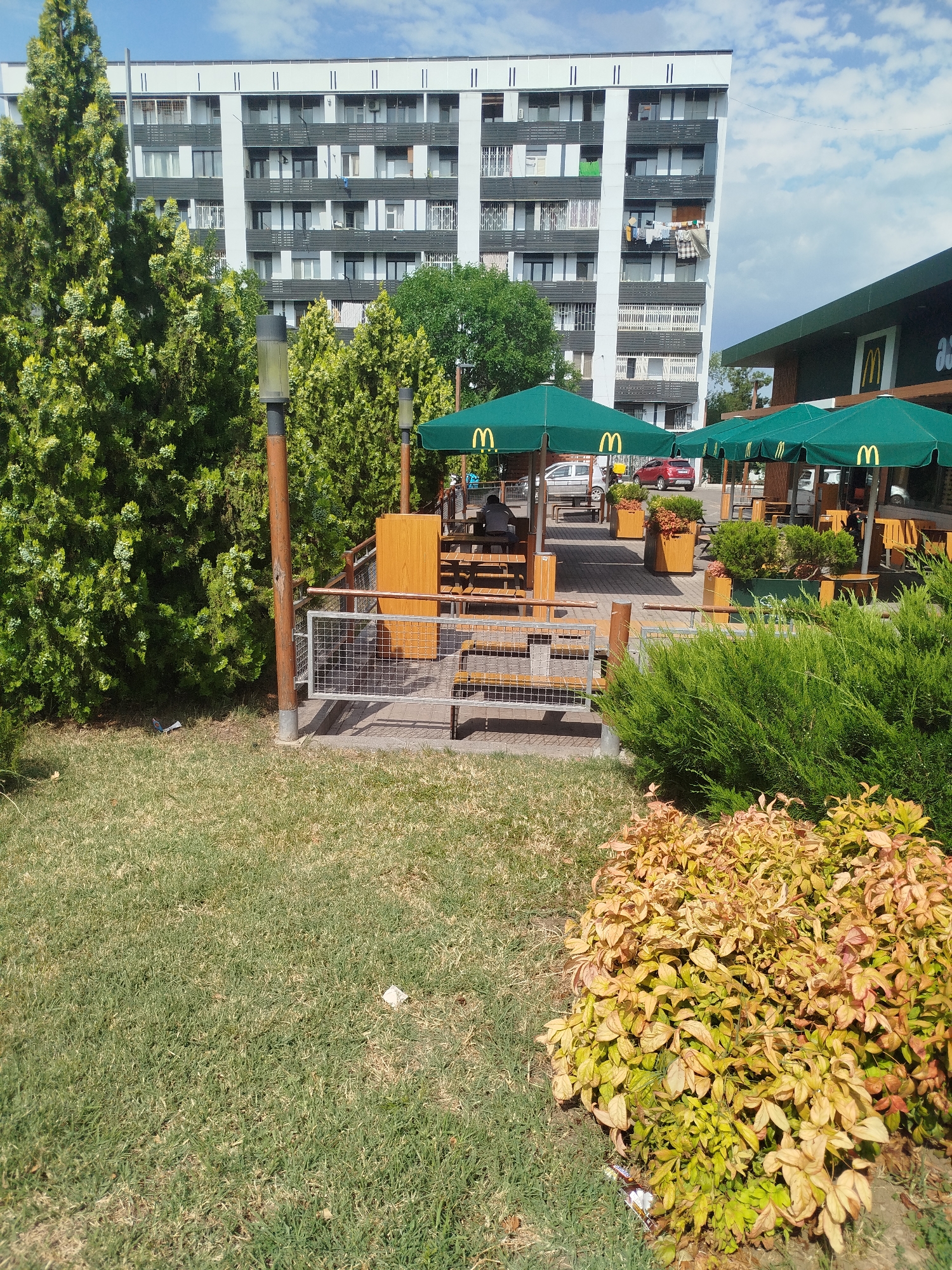 The image shows an outdoor seating area of a McDonald's restaurant in Rustavi, Georgia. The area is furnished with wooden picnic tables and chairs, covered by green McDonald's branded umbrellas. The seating area is paved with brick pavers and surrounded by greenery, including tall trees and bushes. In the background, a large, multi-story apartment building stands tall against a blue sky with scattered clouds. The building has a repeating pattern of windows and balconies, and laundry is visible hanging from some of the balconies, indicating residential use. A portion of the McDonald's restaurant building, with its distinctive green and yellow branding, is visible on the right side of the image. A red car and a white car are parked in the background, near the restaurant. The ground in the foreground is covered with grass, with some patches of dry, yellowed grass and fallen leaves.