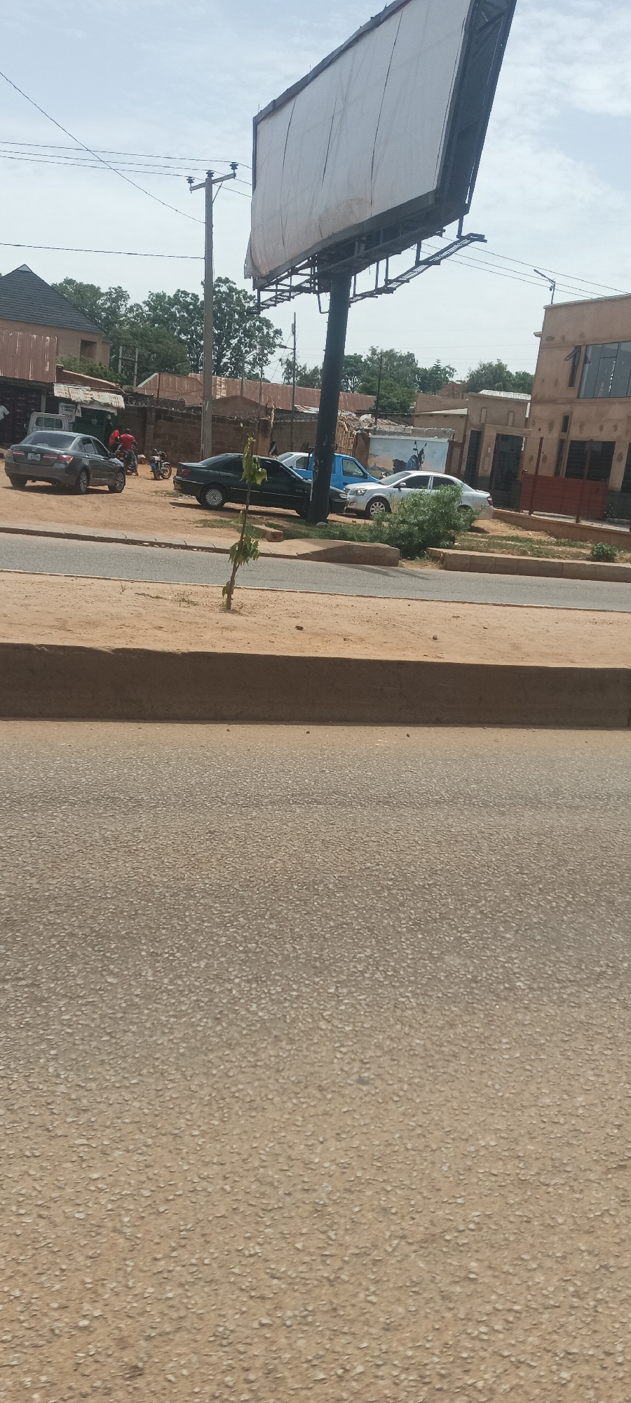 The image shows a street scene in Bukuru, Nigeria. A large blank billboard dominates the upper portion of the frame, supported by a thick, dark pole. Below the billboard and behind the curb line, several parked cars are visible, including a dark sedan, a blue pickup truck, and a white sedan. In the background, people are present, including individuals near a roadside establishment. A power line and utility pole are also visible. The foreground is dominated by a paved road with a dirt shoulder. A small, young tree is growing on the side of the road. The sky is partially overcast.