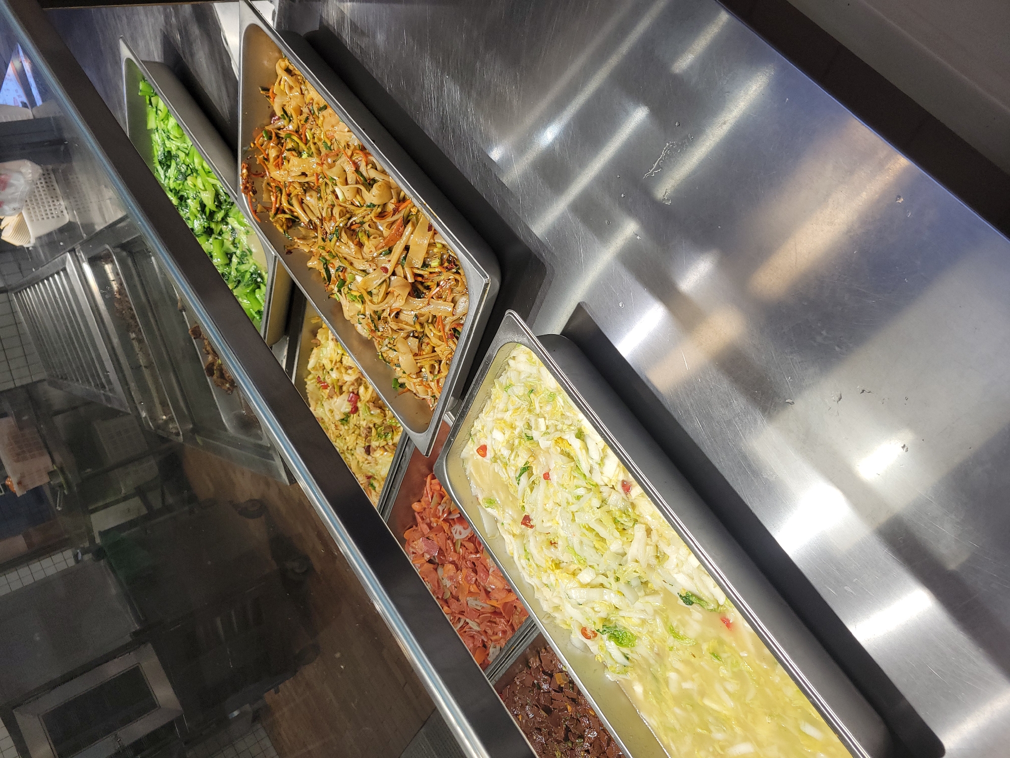 The image shows a food display case, likely located in a restaurant or cafeteria in an unknown city, Unknown. The case contains several metal trays filled with various prepared food items. From left to right, the trays contain green vegetables, a noodle dish with shredded carrots and vegetables, another mixed dish with what appears to be eggs or rice, sliced orange vegetables, and a dish of braised dark brown cubes. The stainless steel case is well-lit and appears clean. No people are visible in the frame. The focus is on the food items within the display case.