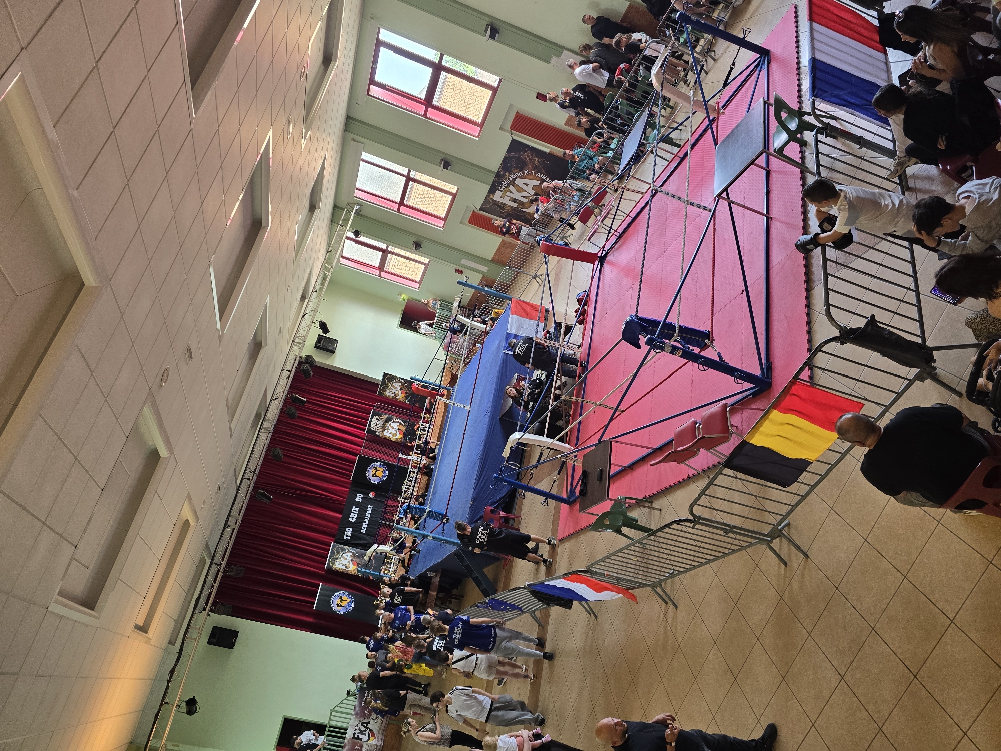 The image depicts an indoor martial arts event held in Berlaimont, France. A boxing ring with a red mat sits in the center, surrounded by spectators. There are flags of France and Belgium displayed around the ring. Banners with the text "THO CHIE DO BERLAIMONT" and "Fédération K-1 Allianc" are visible. The crowd consists of individuals watching the event, some seated, and others standing. A referee wearing a black shirt is present near the ring.
