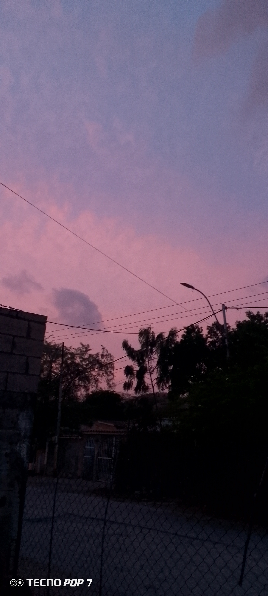 This is a vertical, eye-level, full shot of a twilight sky over a somewhat industrial area. The sky is a gradient of light pink to lavender, with scattered clouds. A single power line runs diagonally across the sky from the upper left to the center. A street lamp on a pole is visible in the upper right quadrant, silhouetted against the sky. In the lower half of the image, there are dark silhouettes of trees and buildings, suggesting they are in shadow. A chain-link fence runs along the bottom of the frame. The lower left corner of the image displays the text "TECNO POP 7."