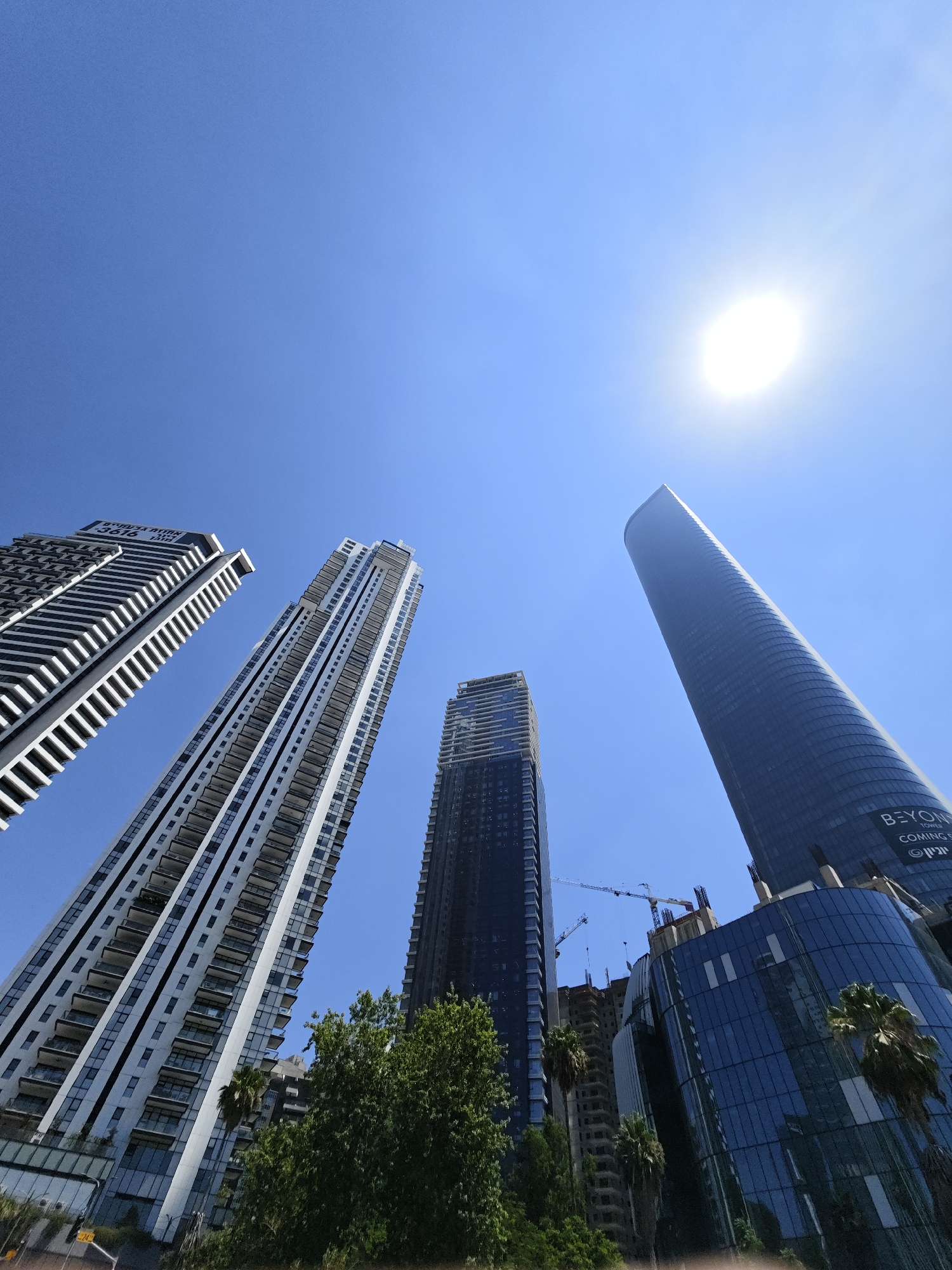 This is a low-angle, wide-shot photograph of several modern skyscrapers under a bright blue sky with the sun high and glinting. The buildings are predominantly made of glass and steel, featuring geometric designs and balconies.
From left to right, the first building is a tall, slender skyscraper with a white facade and dark horizontal bands, its upper sections displaying a repeating pattern of dark geometric shapes. The second building is an even taller, white skyscraper with numerous balconies that are recessed and lined with glass railings. The third building, to the right of the white skyscraper, is a dark, rectangular skyscraper under construction, with visible structural elements and cranes overhead. The fourth and most prominent building on the right is a very tall, cylindrical skyscraper with a dark, reflective glass facade. This building displays text near its base that reads "BEYOND TOWER COMING SOON" and some Hebrew text.
The foreground features lush green trees and palm trees, obscuring the lower parts of the buildings, suggesting a street-level or park perspective looking up. The overall scene conveys an urban environment with ongoing construction and a sense of modern architectural development. The presence of Hebrew text on the cylindrical tower strongly suggests the location is in Israel. Based on the architectural style and the specific text "BEYOND TOWER", this scene is located in Givatayim, Israel, which is known for its high-rise developments.