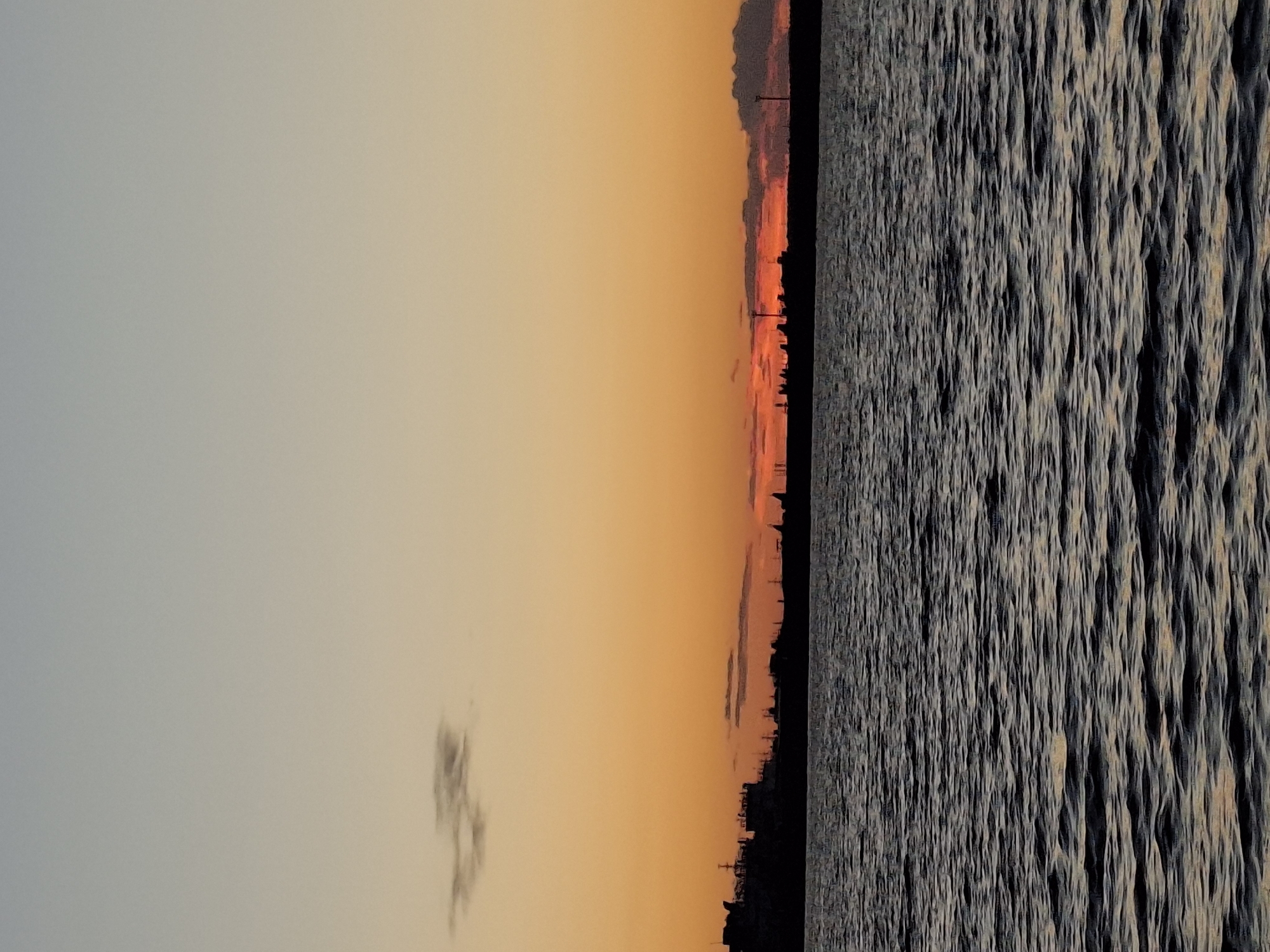 The image depicts a tranquil body of water, likely the Kama River, under a gradient sky of fading twilight hues, transitioning from a pale blue at the top to a warm orange near the horizon. A dark, silhouetted industrial or urban skyline stretches across the middle ground, punctuated by various structures and what appear to be tall towers or chimneys. The water's surface is rippled, reflecting the muted colors of the sky and the dark shapes of the landmass. A single, small, wispy cloud is visible in the upper left quadrant of the sky. The scene conveys a sense of calm and the end of the day.