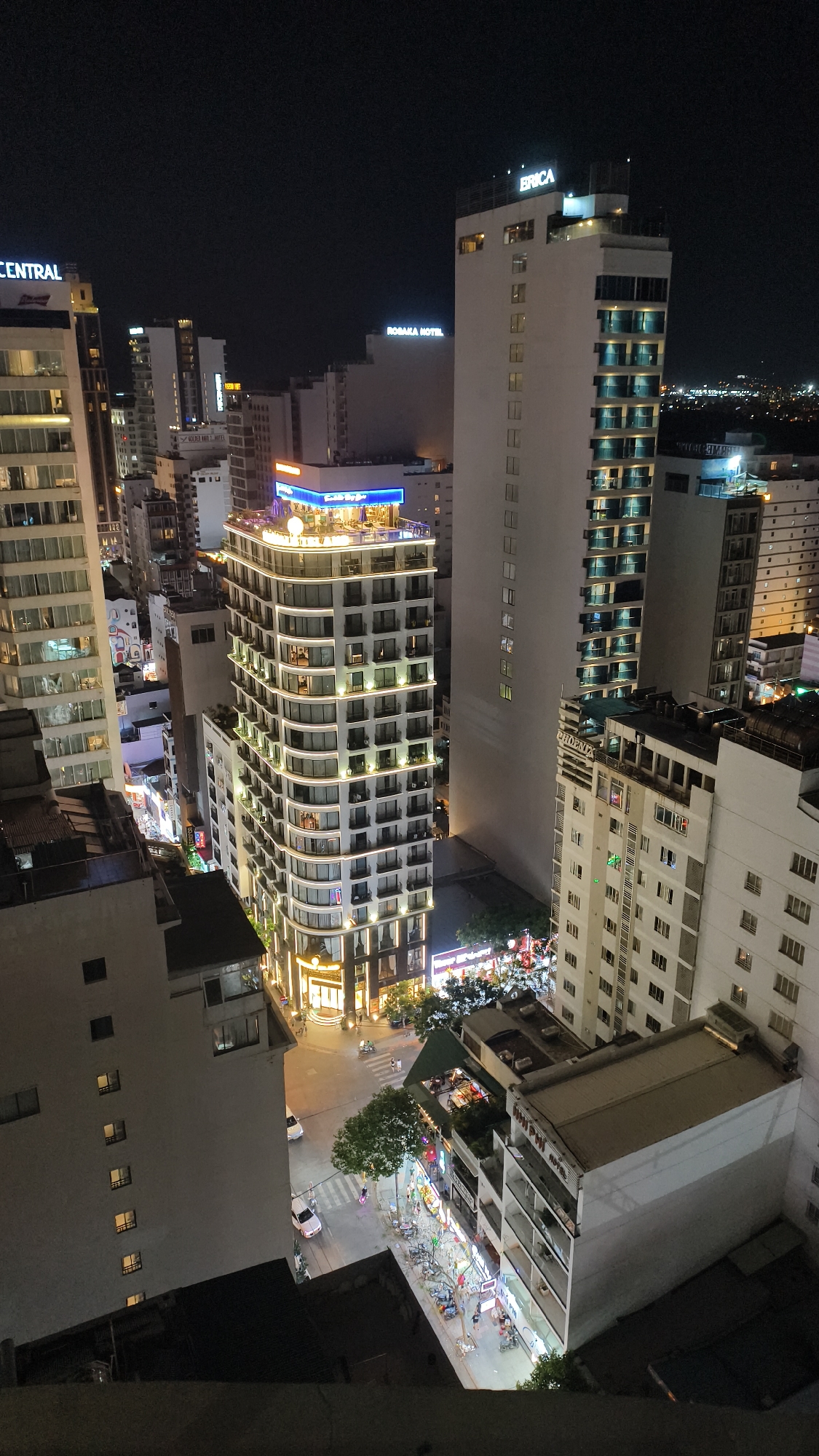 This is a night-time, high-angle shot overlooking a city street scene in Nha Trang Ward, Vietnam. The image captures a dense urban environment characterized by numerous illuminated buildings of varying heights, including hotels and commercial establishments. Prominent signage on buildings identifies "ERICA," "ROSANA HOTEL," and "CENTRAL." The street below is bustling with activity, showing several cars and motorcycles navigating the road, as well as pedestrians walking along the sidewalks. Businesses with brightly lit storefronts and outdoor seating areas are visible, suggesting a vibrant nightlife. Trees and foliage are interspersed among the structures, adding a touch of greenery to the concrete landscape. The overall impression is one of a lively, well-lit urban center at night.