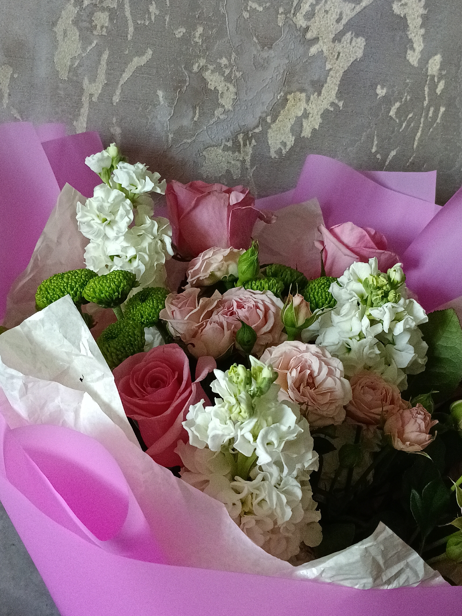A bouquet of flowers is presented in a pink wrapping with white tissue paper accents. The flowers include pink roses of varying shades, white stock flowers, and round green chrysanthemums. The background is a textured gray wall. The location is Chaikovsky, Russia.