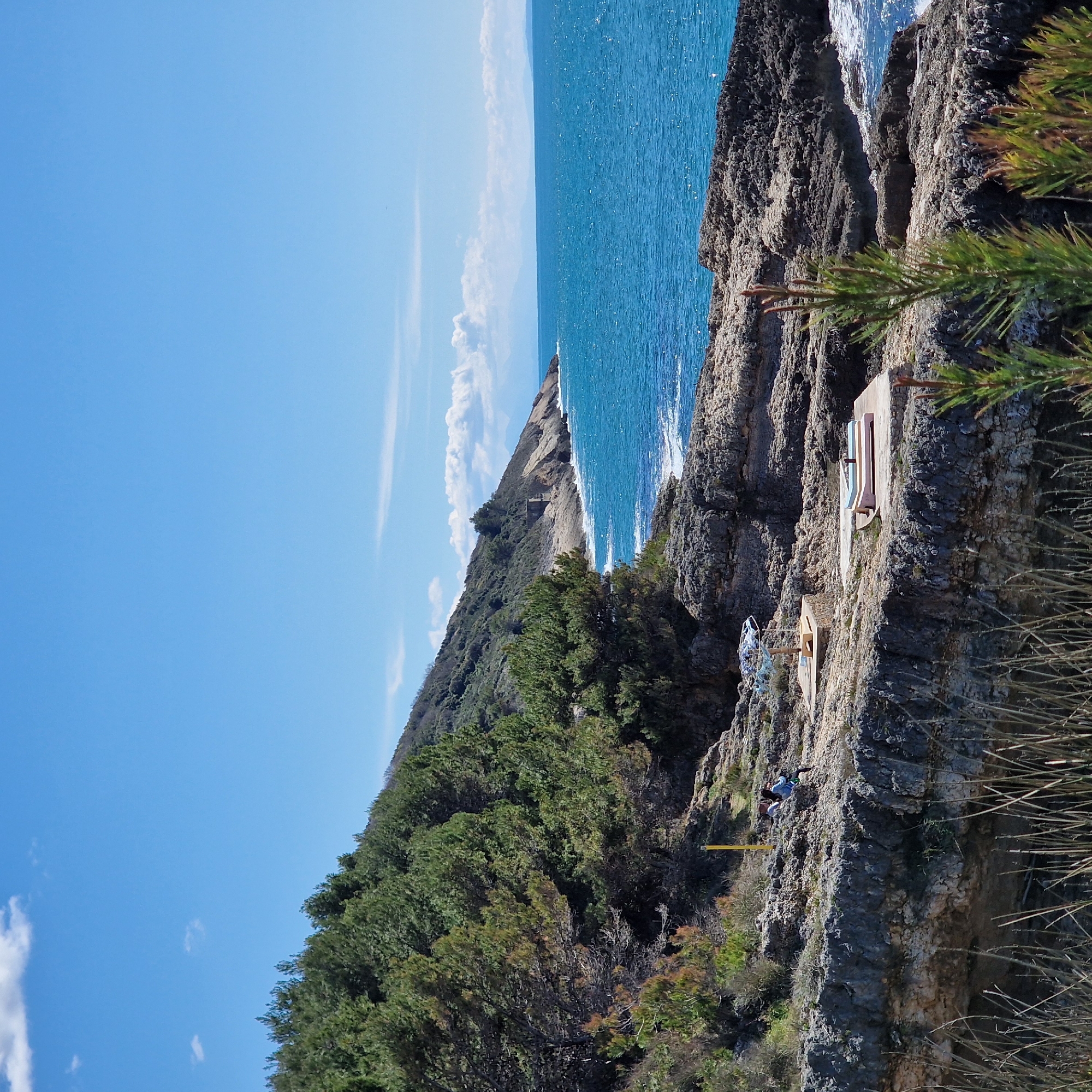 The image depicts a coastal scene in Ulcinj - Ulqin, Montenegro. The Adriatic Sea occupies the top right, a clear blue contrasting with rocky cliffs. A forested hillside rises on the left, sloping down to meet the sea. Structures, possibly remnants of fortifications or lookout points, cling to the cliffside. A person or persons are visible on the cliff edge. The sky is a clear blue with scattered white clouds.