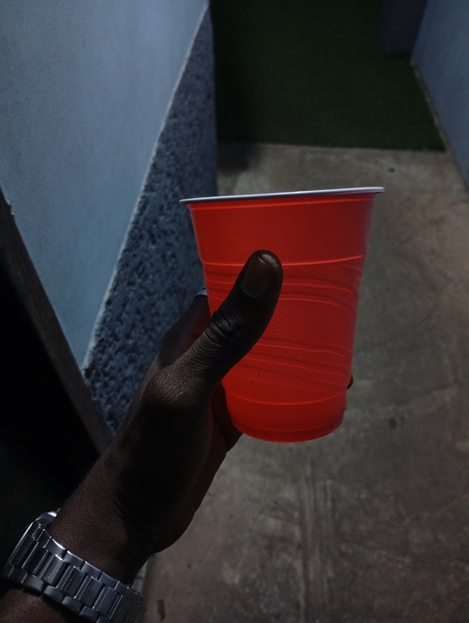 A person holds a red plastic cup in their right hand. The person is wearing a silver wristwatch on their left wrist. The background consists of a wall with a textured surface to the left, a paved walkway or patio in the center, and a patch of green vegetation in the upper right. The scene appears to be outdoors and is dimly lit. It is in Adankolo, Nigeria.