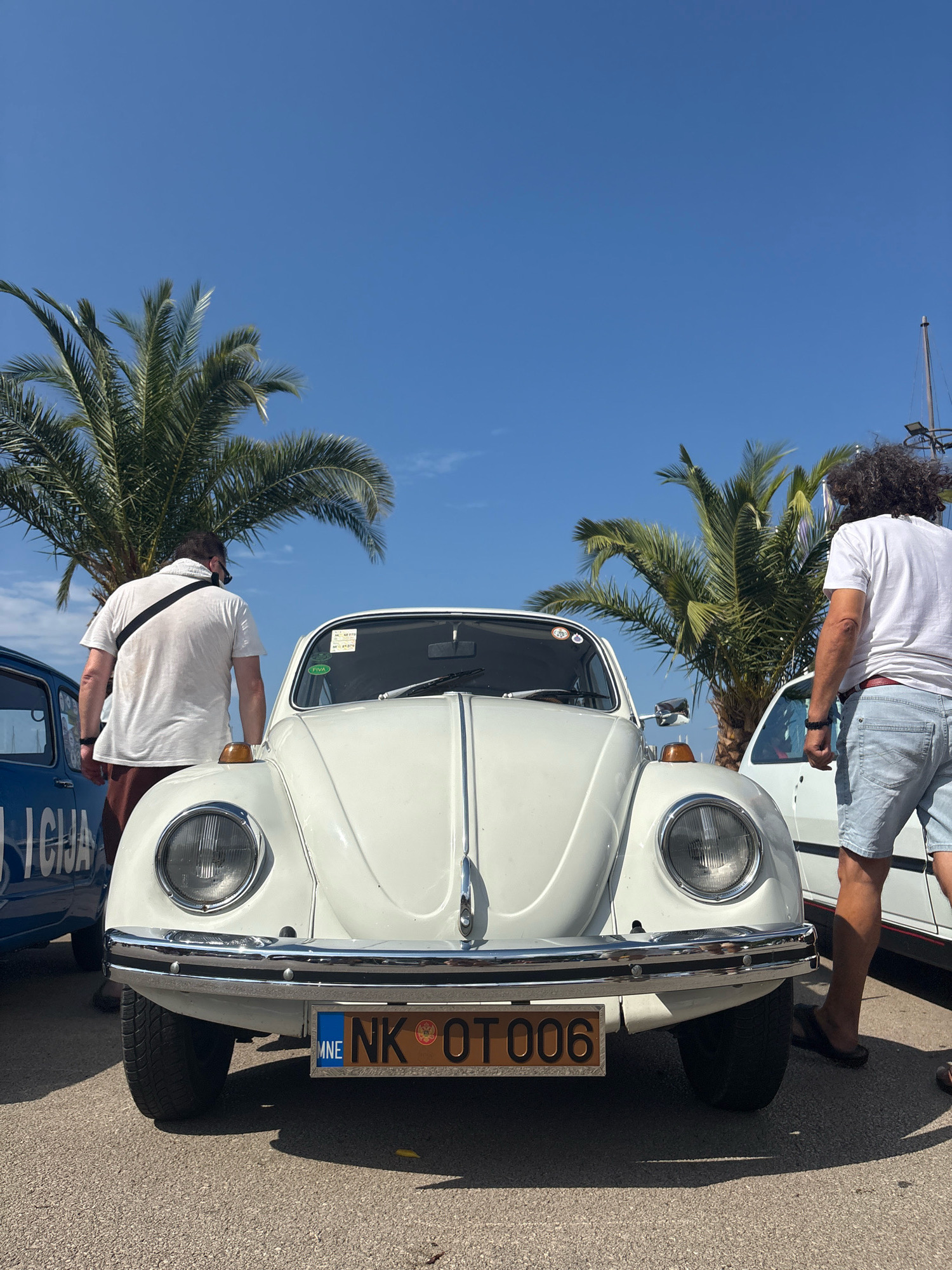 The image captures a white Volkswagen Beetle parked outdoors on a sunny day, likely in the city of Bar, Montenegro. The car is the central subject, viewed from the front. It has round headlights, a chrome bumper, and a license plate reading "MNE NK 0T006". To the left of the Beetle, a portion of a blue car with text visible on its side can be seen. Two men are present in the scene. One man, wearing a white t-shirt and dark shorts, stands behind the Beetle, facing away from the camera. The other man, wearing a white t-shirt and light blue denim shorts, stands to the right of the Beetle, also facing away. Both men appear to be observing the cars. Palm trees are visible in the background, and the sky is a clear blue, indicating good weather. The ground appears to be asphalt. The overall scene suggests an outdoor car gathering or display.