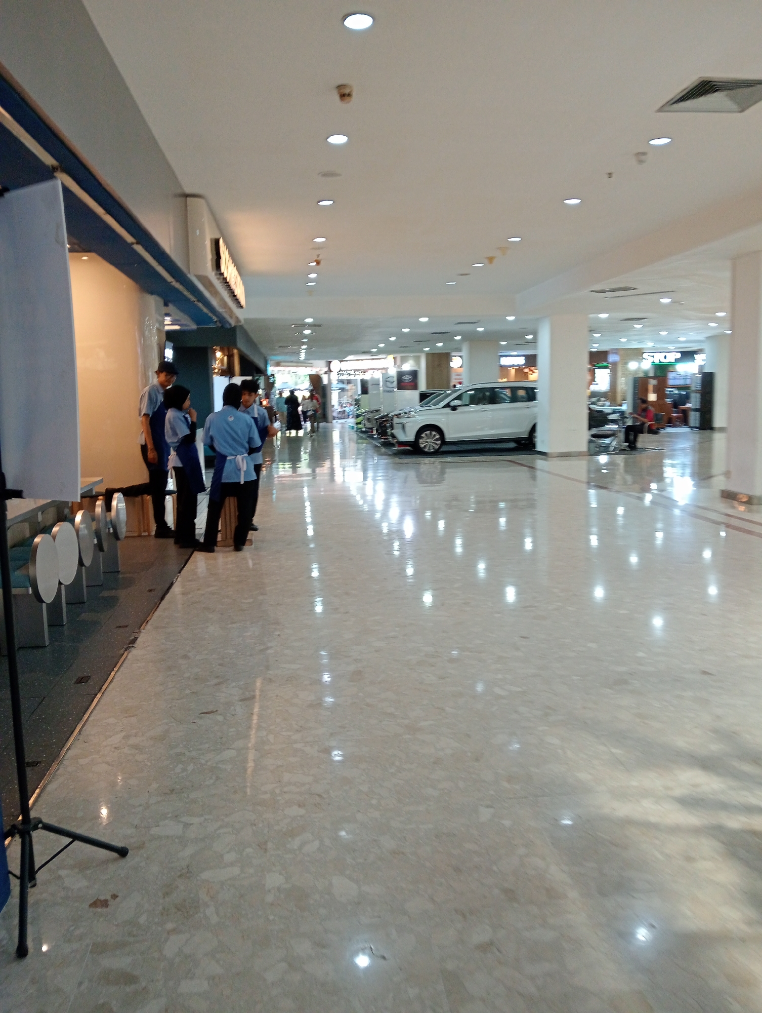 The image captures a wide-angle view of an indoor commercial space, likely a shopping mall or exhibition hall, in Surabaya, Indonesia. The scene is brightly lit by overhead lights, reflecting off the polished marble floor. In the foreground on the left, a portion of a black tripod and a blue object are visible. To the right of this, a group of four individuals are standing near a display area. They appear to be employees, wearing matching blue shirts with white aprons or smocks, and dark pants. One person is wearing a dark cap. They are facing towards the center of the space and seem to be engaged in a conversation or receiving instructions.
Further into the background, a white car is prominently displayed on the right side of the walkway, suggesting a car exhibition or a dealership within the mall. Beyond the car, several shops or food stalls are visible, one with a sign that reads "STOP". The architecture of the space is modern, with a suspended ceiling and columns. People can be seen walking in the distance, indicating general public activity within the establishment. The overall impression is one of a clean, well-maintained, and busy commercial environment.