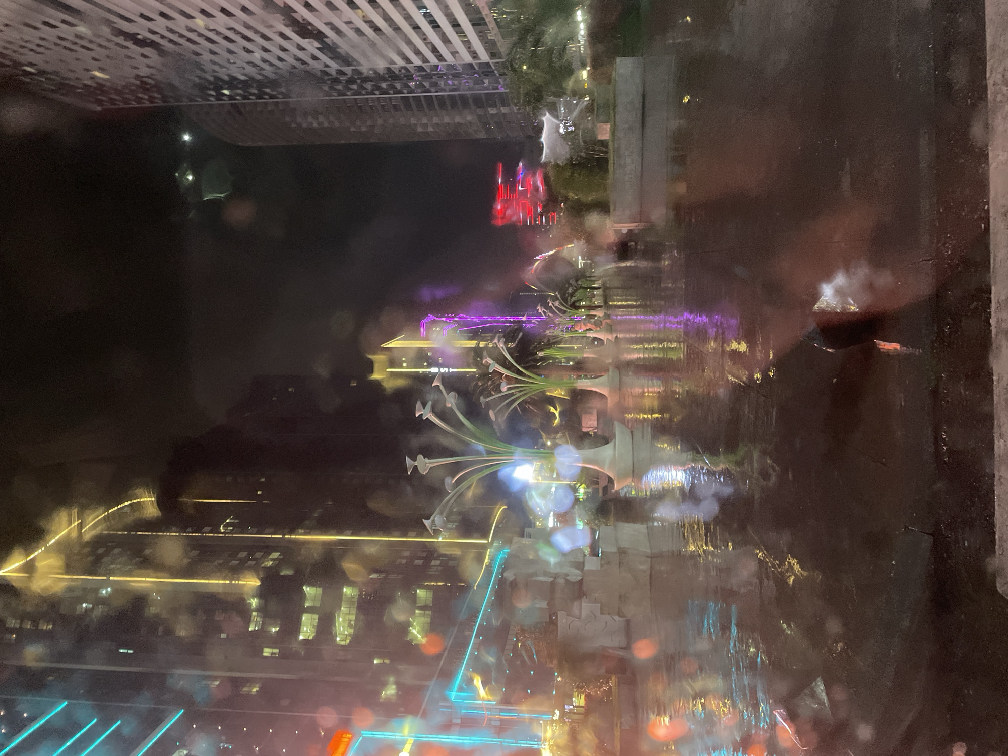 This appears to be a nighttime photograph taken through a wet or rain-streaked window, showing a view of the city of Batumi, Georgia. The image is oriented sideways. There are multiple buildings with lit windows visible in the background. There are some bright and colorful neon lights from a large monument or artwork on the left side, along with water features. There is a prominent sculpture in the center of the frame. In the upper portion, there is another building, and a red light source. The photograph seems to capture the reflections and distortions caused by rain or moisture on the window. The environment is a city at night.