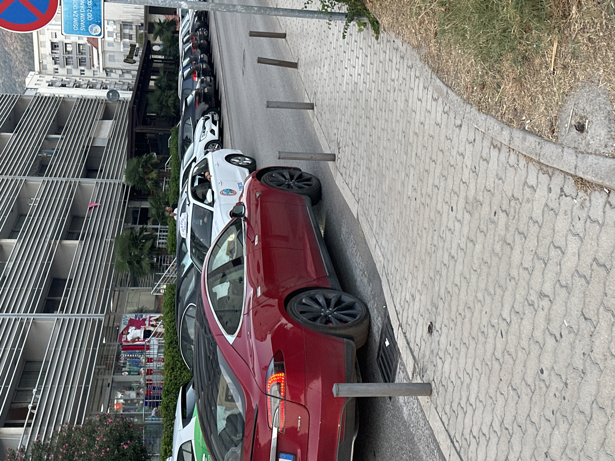A dark red Tesla Model S is parked on the left side of a street in Budva, Montenegro, viewed from the rear passenger side. The car's taillights are illuminated. In the background, a row of parked cars, including white taxis, are visible along the street. A pedestrian walkway with gray brick pavers runs along the right side of the street, separated by bollards. Behind the walkway is dry grass and a curb. A multi-story building with horizontal balconies and a storefront is located behind the parked cars. A traffic sign is visible on the right side of the image.