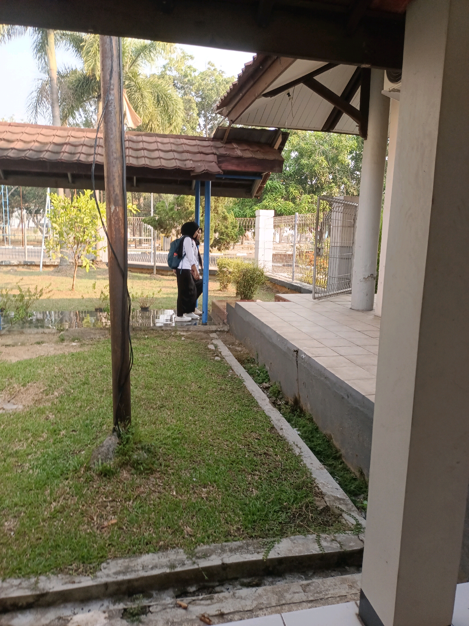 The image depicts an outdoor scene with two individuals standing near a covered walkway. The environment appears to be a public space, possibly part of a school or community area, with a grassy lawn, a tiled pathway, and a metal fence in the background. Palm trees and other foliage are visible, suggesting a tropical or subtropical climate. One person is wearing a white shirt and black pants, with a backpack on, while the other is wearing dark clothing. They are standing under a roofed structure with tiled roofing. The ground is a mix of grass and some paved areas. A prominent utility pole stands in the foreground. The lighting suggests it is daytime, possibly in the morning or afternoon, with the sun casting shadows.