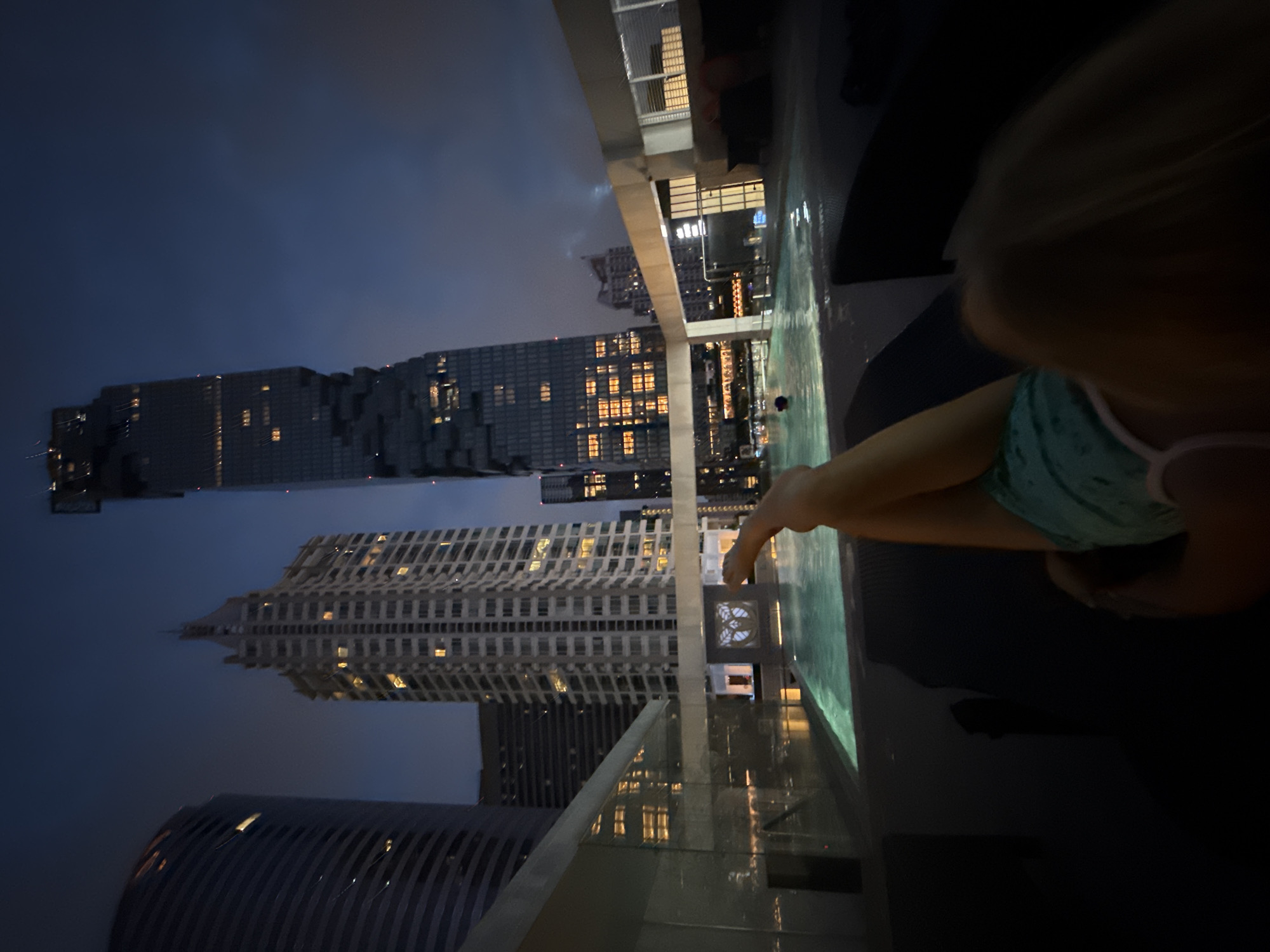 In Bangkok, Thailand, at dusk, a person's legs are visible from the knees down, resting on a dark lounge chair. The person is wearing teal patterned shorts. In the foreground, a rooftop swimming pool is illuminated by turquoise lights. In the background, several skyscrapers pierce the twilight sky, their windows aglow with artificial light. The most prominent skyscraper, the MahaNakhon building, features a distinctive pixelated facade. The overall atmosphere is one of urban leisure and nighttime city life.