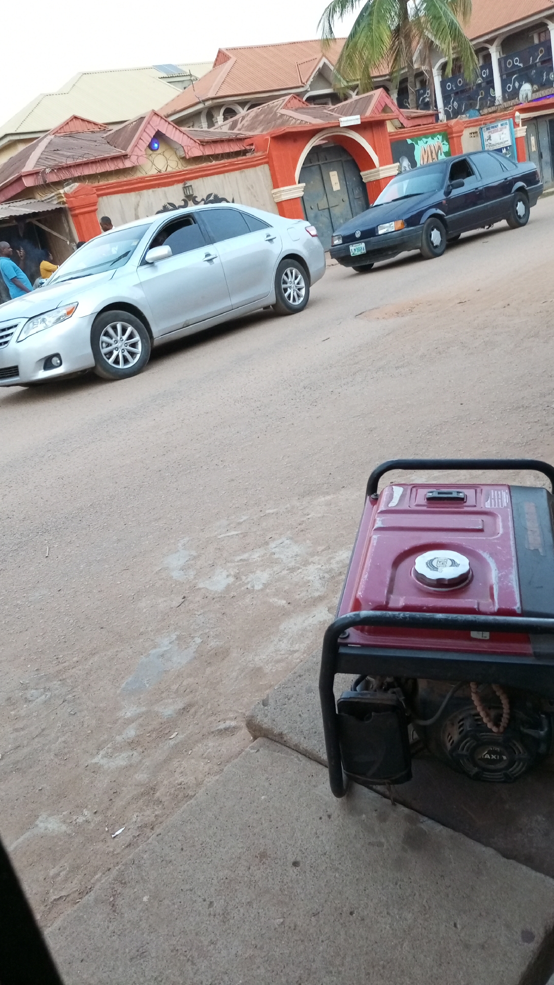 The image captures a street scene in Sabon Tasha, Nigeria. A silver Toyota Camry and a dark-colored Volkswagen Passat are parked diagonally on the road. The architecture visible in the background includes buildings with red-tiled roofs. In the foreground, a red and black portable generator rests on a concrete surface. A partial view of people can be seen near a building in the background. The road surface appears unpaved and dusty.