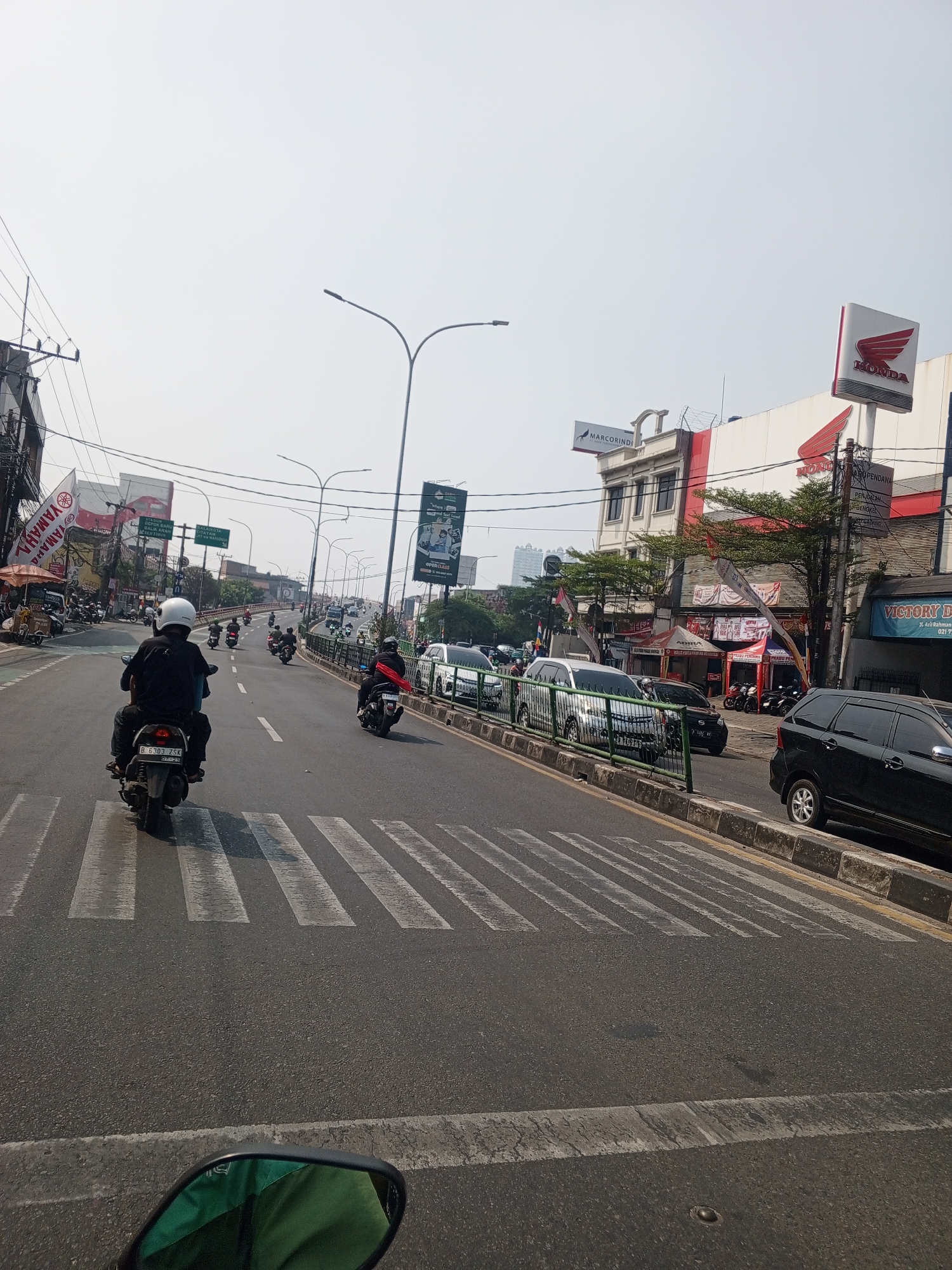 The image captures a street scene in Depok, Indonesia, on a bright, hazy day. In the foreground, a motorcycle with two occupants is in the process of crossing a pedestrian crossing. The driver, wearing a black shirt and helmet, is in control, while the passenger is seated behind. The road is paved and shows signs of wear, with distinct white stripes marking the pedestrian crossing.
Further down the road, several other motorcycles and cars are visible, indicating moderate traffic flow. Some motorcycles are parked on the side of the road, and a few are in motion, following the general direction of the main traffic. On the right side of the road, there is a row of shops and buildings. One prominent building features a large white sign with the red "HONDA" logo, suggesting a car dealership. Adjacent to it, another building has a sign that reads "VICTORY D....". Overhead, utility poles and wires are visible, typical of urban infrastructure.
On the left side of the road, there are more shops and advertisements, including a large "YAMAHA" banner. Traffic lights and street lamps are also present, lining the road and sidewalk. The overall environment appears to be a busy commercial and traffic artery within the city. The sky is clear but appears washed out, possibly due to atmospheric conditions or the camera's exposure. The scene is consistent with a typical urban street in Indonesia during daylight hours.