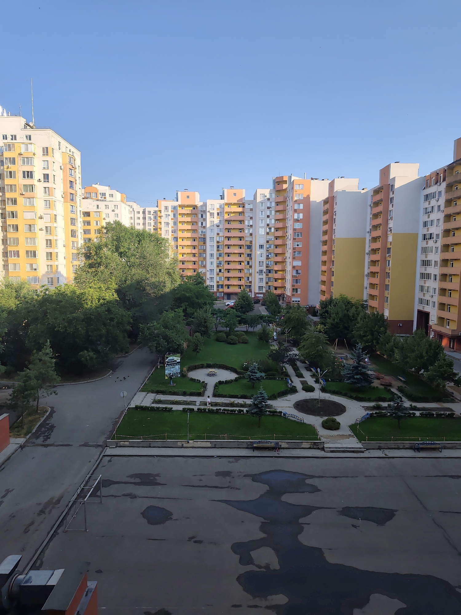 The image shows a residential area with tall apartment buildings in Chișinău, Moldova. The buildings are multi-story structures with balconies and windows. In the foreground, there is a paved courtyard with puddles of water, possibly from recent rain. A soccer goal frame is visible in the courtyard. Behind the courtyard, there is a green park area with trees, benches, and walking paths. A playground with swings and slides is situated within the park. Several people are walking in the park. The sky is clear and blue. The scene appears to be an urban residential setting.