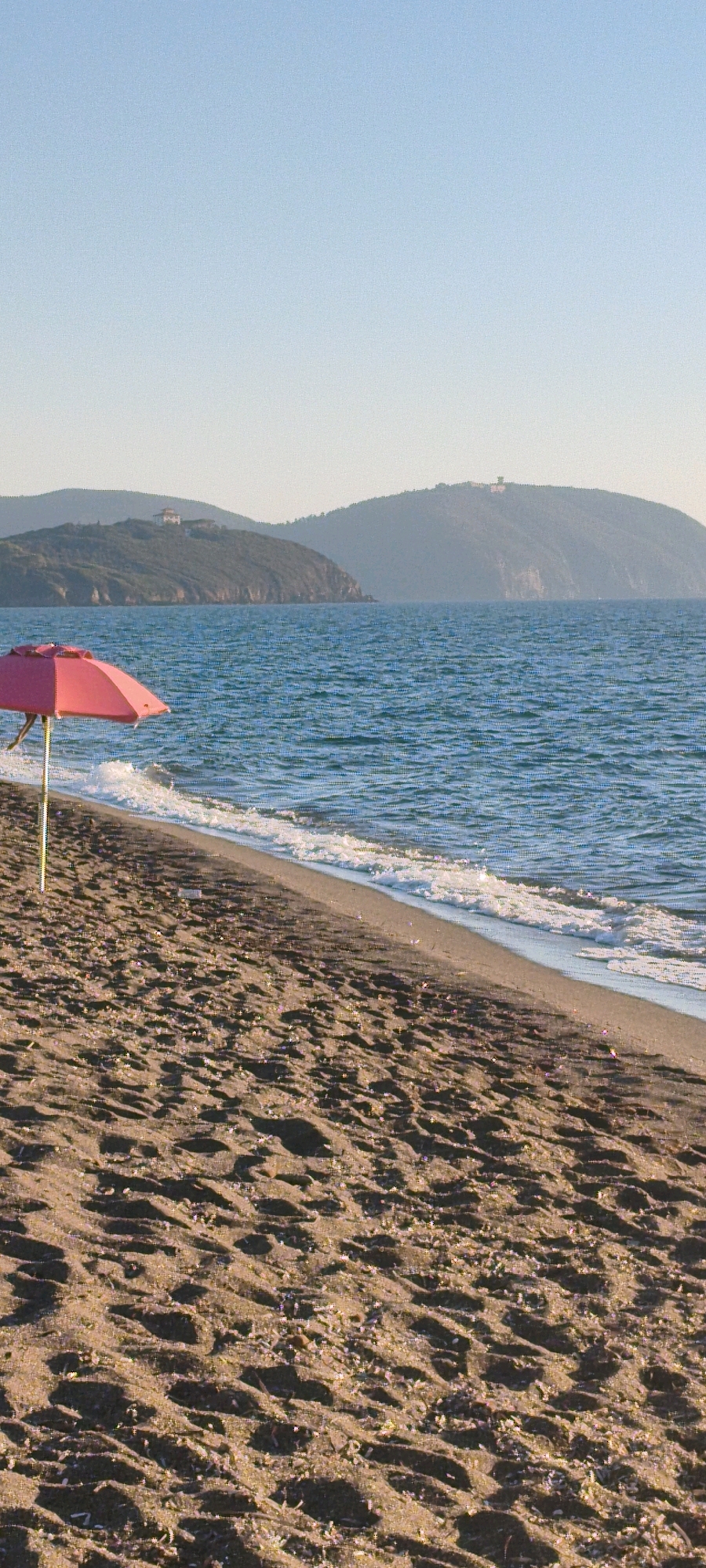 A low-angle, vertical medium shot captures a peaceful beach scene in San Vincenzo, Italy, under a clear, pale blue sky. In the foreground, the sandy beach is textured with shadows and scattered small, light-colored objects, possibly shells or pebbles. A bright pink beach umbrella, its fabric slightly creased, stands open on a thin yellow pole, positioned on the left side of the frame, close to the water's edge. The sea occupies the middle ground, its surface a deep blue with subtle ripples and white foam marking the gentle waves lapping onto the shore. Beyond the sea, rolling, tree-covered hills form the background. A small building with a white roof is visible on the crest of the furthest hill. The overall atmosphere is tranquil and serene.