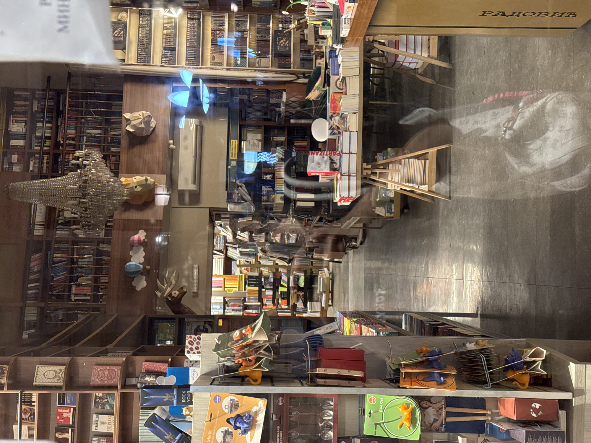 The image depicts the bookstore "Geca Kon," located in Belgrade, Serbia. The view is from outside, looking in through a glass window, which causes a reflection of the external environment. Inside, the bookstore is filled with wooden shelves stacked high with books. The bookstore features chandeliers and wooden animal sculptures for decoration. There is also a display of smaller items on a shelf near the front, including games, books, and other small objects. The bookstore's name is visible on a wooden sign above the shelves.