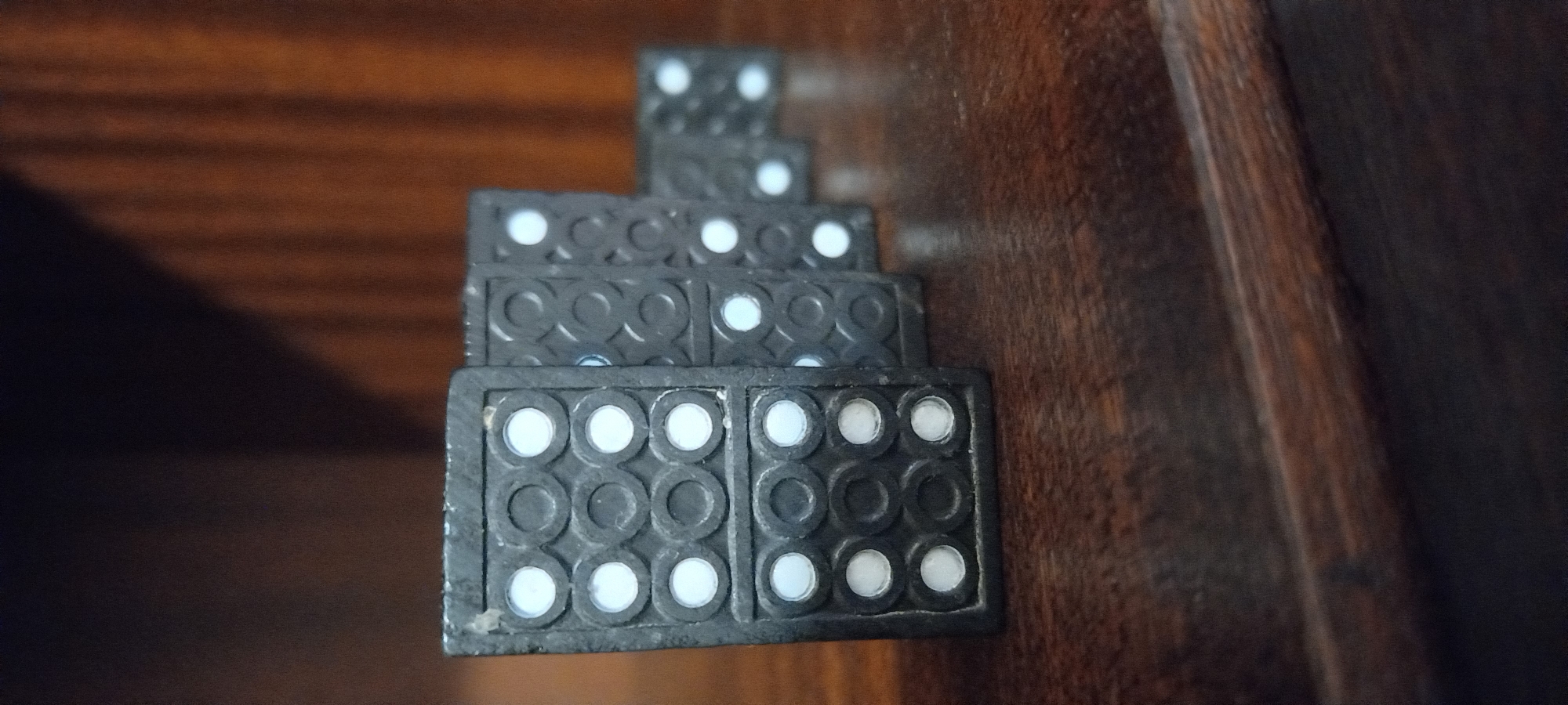 This close-up image features a stack of five black dominoes with white pips, arranged in a receding, stair-step formation against a warm-toned wooden background. The dominoes appear to be old, made of a dark material with circular indentations surrounding the white pips.
In the foreground, the largest domino is clearly a double-nine, with a 3x3 arrangement of white pips on each half. Behind it, a double-seven domino is partially visible, followed by what appears to be a double-five or double-six. Further back are a double-four and, at the top of the stack, a double-two, which is somewhat out of focus. Some of the white pips show signs of wear or slight discoloration.
The setting is an indoor environment, indicated by the visible wood grain of what appears to be furniture—perhaps a table, shelf, or cabinet. The wooden surface displays varying shades of brown. A significant dark shadow is cast on the lower left portion of the background, while a darker, possibly curved, wooden edge runs along the right side of the frame.
There are no people or discernible activities taking place; the dominoes are stationary. No specific time of day, weather conditions, or emotions are conveyed. There is no visible text in the image. The scene is a still-life, focusing on the texture, material, and arrangement of the dominoes.