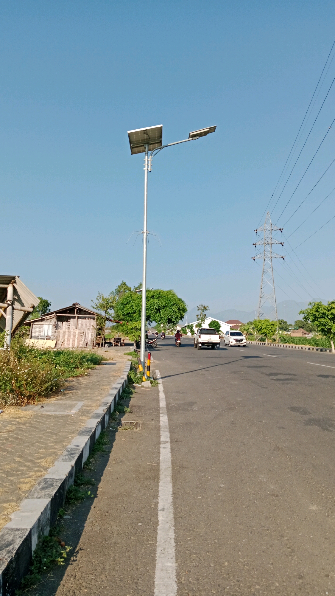 A vertical, eye-level, full outdoor shot captures a road in Sukowinangun, Indonesia, under a clear blue sky. A solar-powered street lamp with two lights stands prominently on the left, its panels angled towards the sun. To its left, a sidewalk runs along the edge of the road, bordered by a curb. Beyond the sidewalk, a rustic wooden structure, possibly a shack or small building, is visible, with overgrown vegetation in front of it.
The road itself is asphalt, marked with a white lane divider. Several vehicles are present: a white pickup truck and a white car are driving on the road, and a motorcycle with a rider is also visible. In the background, a large electricity pylon with multiple wires stretches across the frame, and lush green trees dot the landscape. Distant hills or mountains are faintly discernible against the horizon. The overall impression is that of a rural or semi-rural roadside scene on a sunny day.