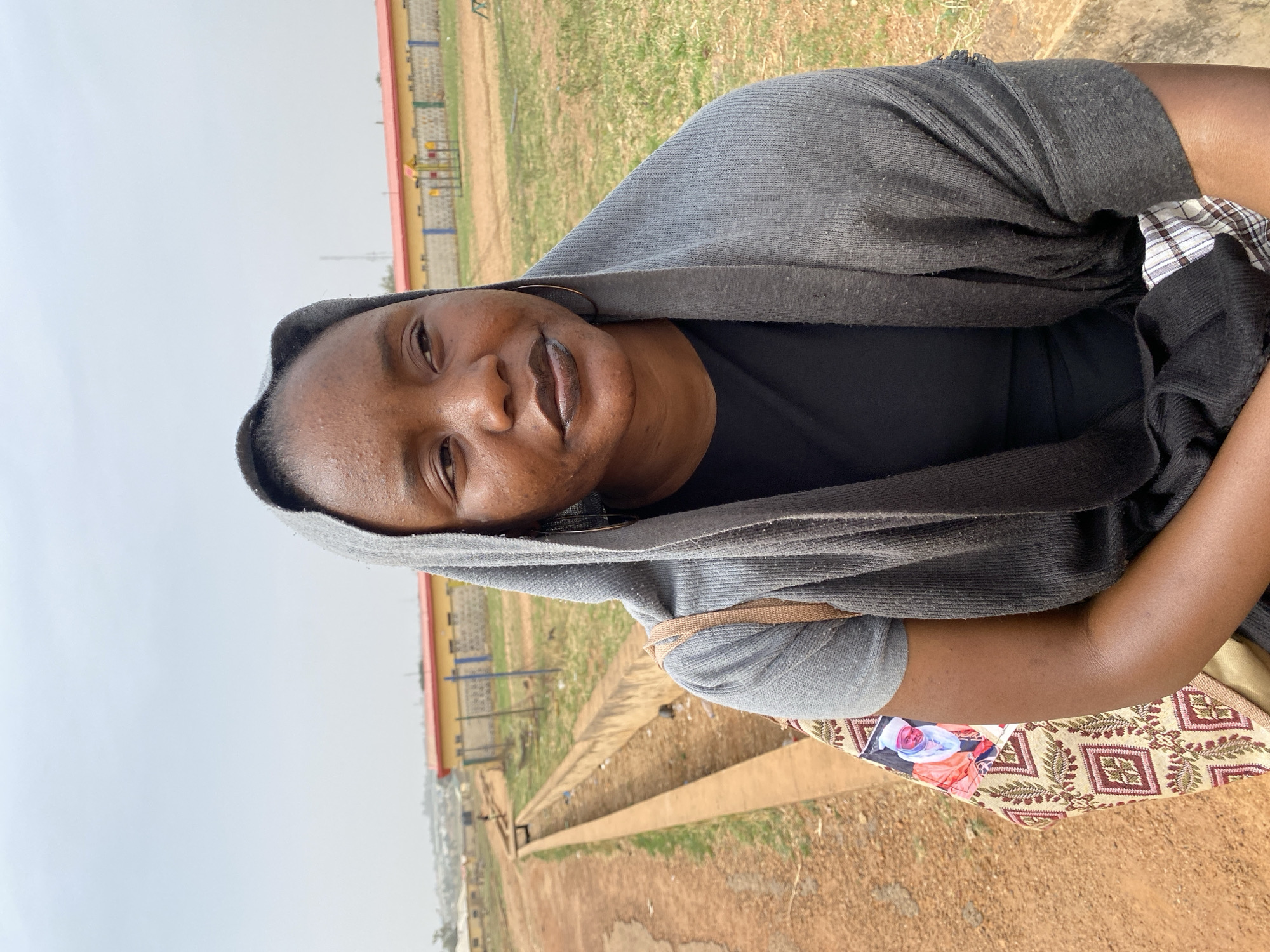 An adult individual, likely female, with dark skin, is captured in an outdoor portrait, smiling softly and looking directly at the camera. The person wears a dark grey hooded top or wrap over a black t-shirt, with a lighter grey long-sleeved garment visible on one arm. A single hoop earring is seen in the visible ear. Their expression is pleasant and calm.
The setting appears to be a dry, open field with sparse green and brown grass under an overcast or hazy sky. A concrete drainage channel or ditch runs parallel to the foreground, separating the viewer from the distant background. In the mid-ground, there's a building with a yellow wall and a red roof, along with colorful playground equipment behind a low fence or wall. Distant structures or hills are faintly visible on the horizon beneath the hazy sky, suggesting an expansive outdoor area.
The individual is holding a patterned fabric bag with a geometric design in beige, red, and brown tones. A printed image of a person, possibly a child in traditional attire (white head covering and orange/pink garment), is visible on the bag. A light brown strap, possibly from another bag, is visible on the person's shoulder. The image appears to have been taken during the daytime, likely in Zawan, Nigeria, given the contextual information. No discernible text is visible in the frame.