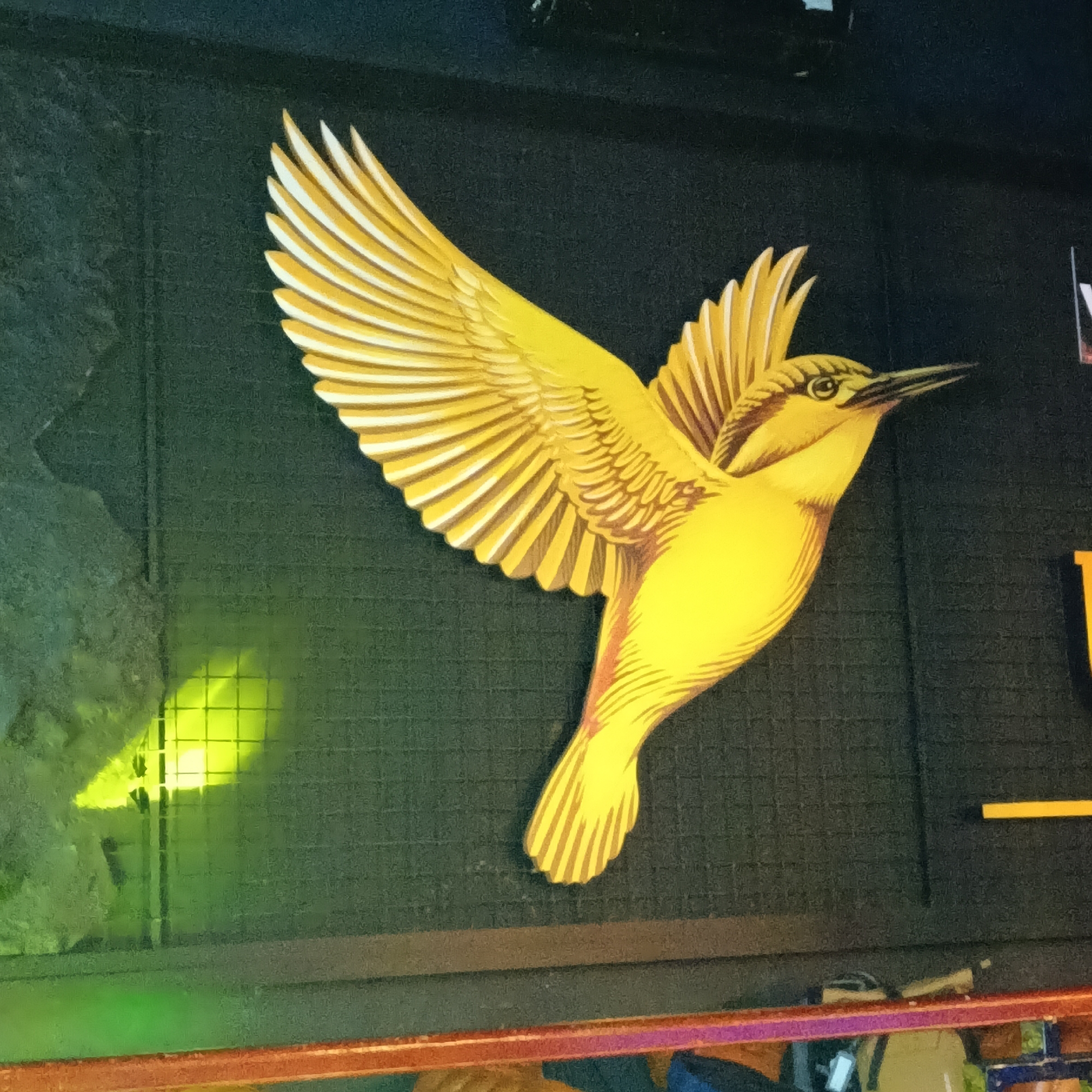 A golden bird, possibly an oriole, is affixed to a dark wall. The bird has detailed wings with individual feathers delineated, and its body is a solid yellow color with some brown shading around the wing joint. The wall has a textured surface, and there is a blurred green object with a grid pattern to the left of the bird. Some architectural details are visible in the lower portion of the frame. Based on the prompt, this is located in Sinquerim, India.