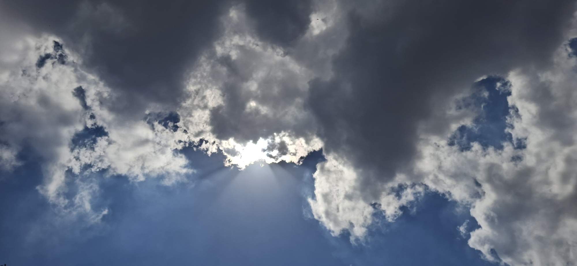The image captures a scene of clouds in the sky, possibly taken in Vallecito, Venezuela, as indicated in the prompt. Predominantly, the view is filled with a dynamic cloudscape, displaying a mix of light and dark clouds. Light from the sun is visible, breaking through a gap in the clouds, creating a ray effect. The sky visible through the clouds is blue. No people, subjects, or objects are discernible besides the clouds and the sky. There are no actions or interactions depicted.