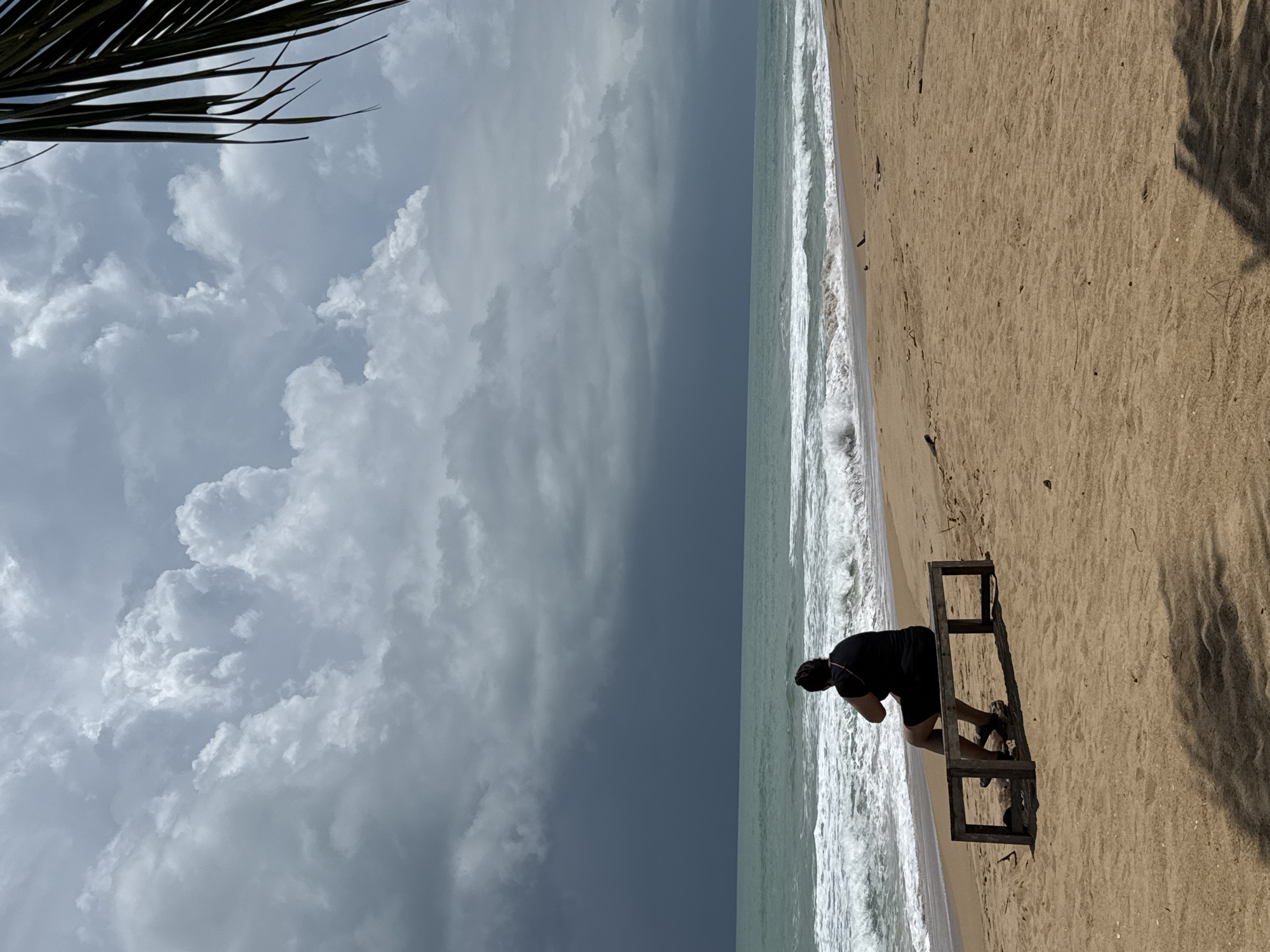 A person is sitting on a wooden bench on a sandy beach, facing the ocean. The sky is dramatic with dark, heavy clouds approaching from the left, contrasting with lighter, puffy clouds on the right. The ocean is a turquoise color with white waves breaking on the shore. A palm frond is visible in the upper right corner of the image. The beach is mostly empty, with the exception of the person on the bench and some scattered debris. The scene suggests an approaching storm. The location is the beach in Ban Khuk Khak, Thailand.
