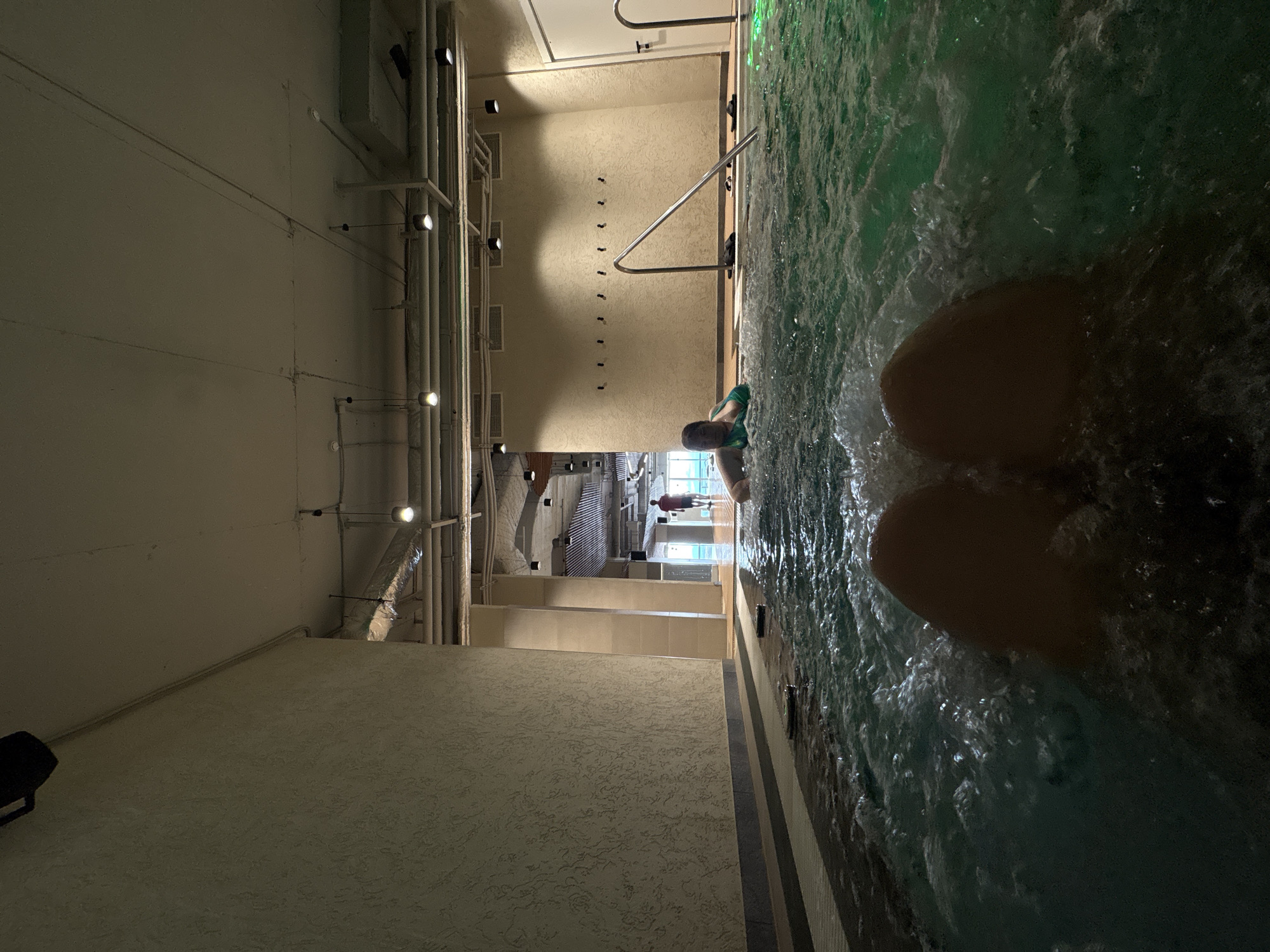 A first-person perspective shot shows a person's legs submerged in a bubbling, greenish hot tub. A woman in a green swimsuit is visible partially submerged in the water on the left side of the frame. The hot tub is indoors, with textured walls and a ceiling with exposed pipes and light fixtures. In the background, a person in a red shirt walks away from the camera towards a brightly lit area with large windows. There are several hooks on the wall behind the woman, and a metal railing is visible in the hot tub. The scene is illuminated by artificial lights. No specific details are provided to identify the location as Khabarovsk, Russia.