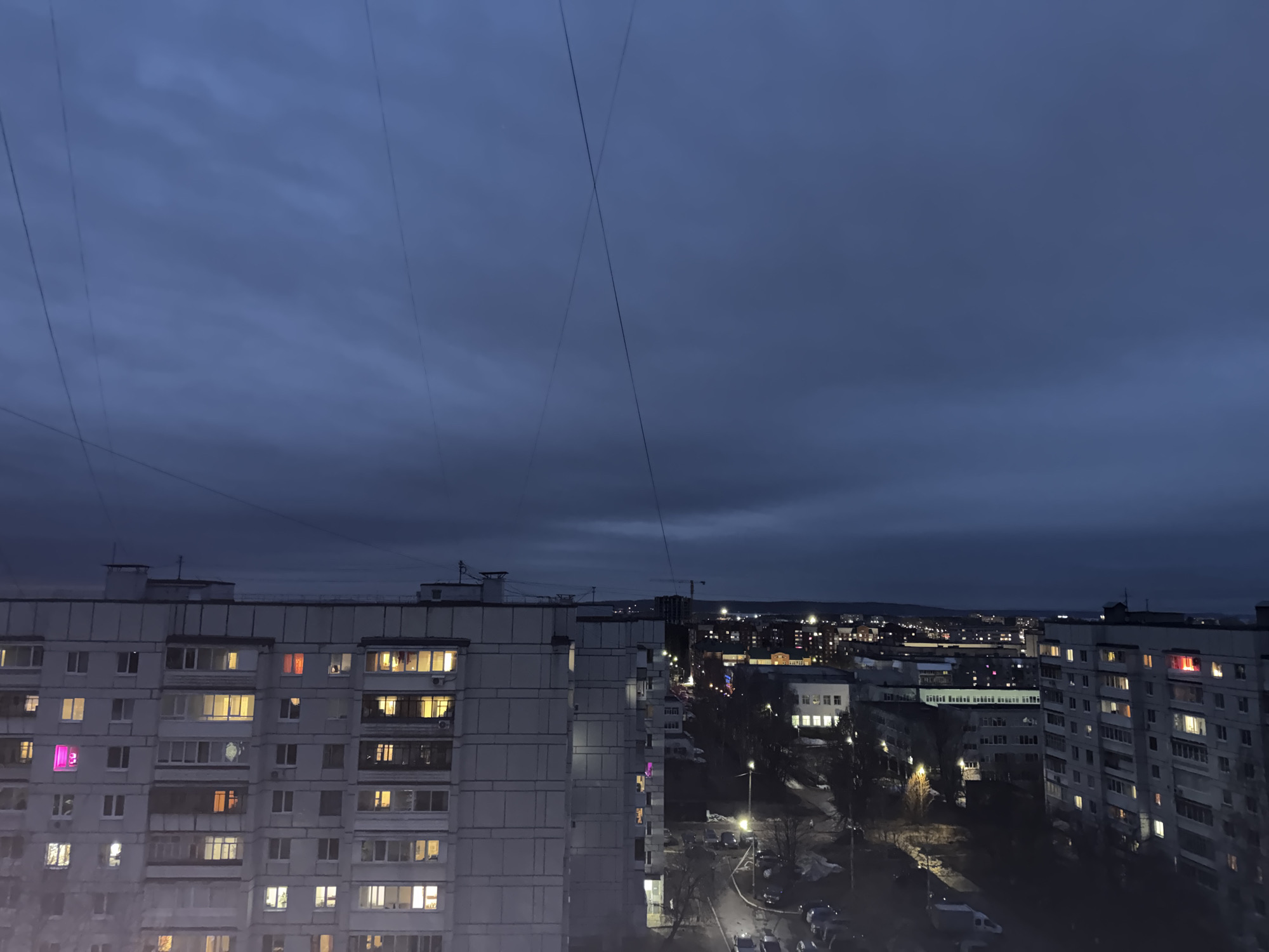 The image shows a night view over the city of Chaikovsky, Russia, from a high vantage point. The foreground is dominated by several large, Soviet-era apartment buildings, the surfaces are gray and monolithic. Some windows are lit with warm, yellow light, while others are dark. Some windows show a purple hue. In the middle ground, a street is visible, illuminated by streetlights. Parked cars line the street. In the background, the cityscape extends into the distance, with numerous lights dotting the horizon. The sky is dark and cloudy. A crane is visible among the city lights. Several thin wires span the sky from the upper left of the image.