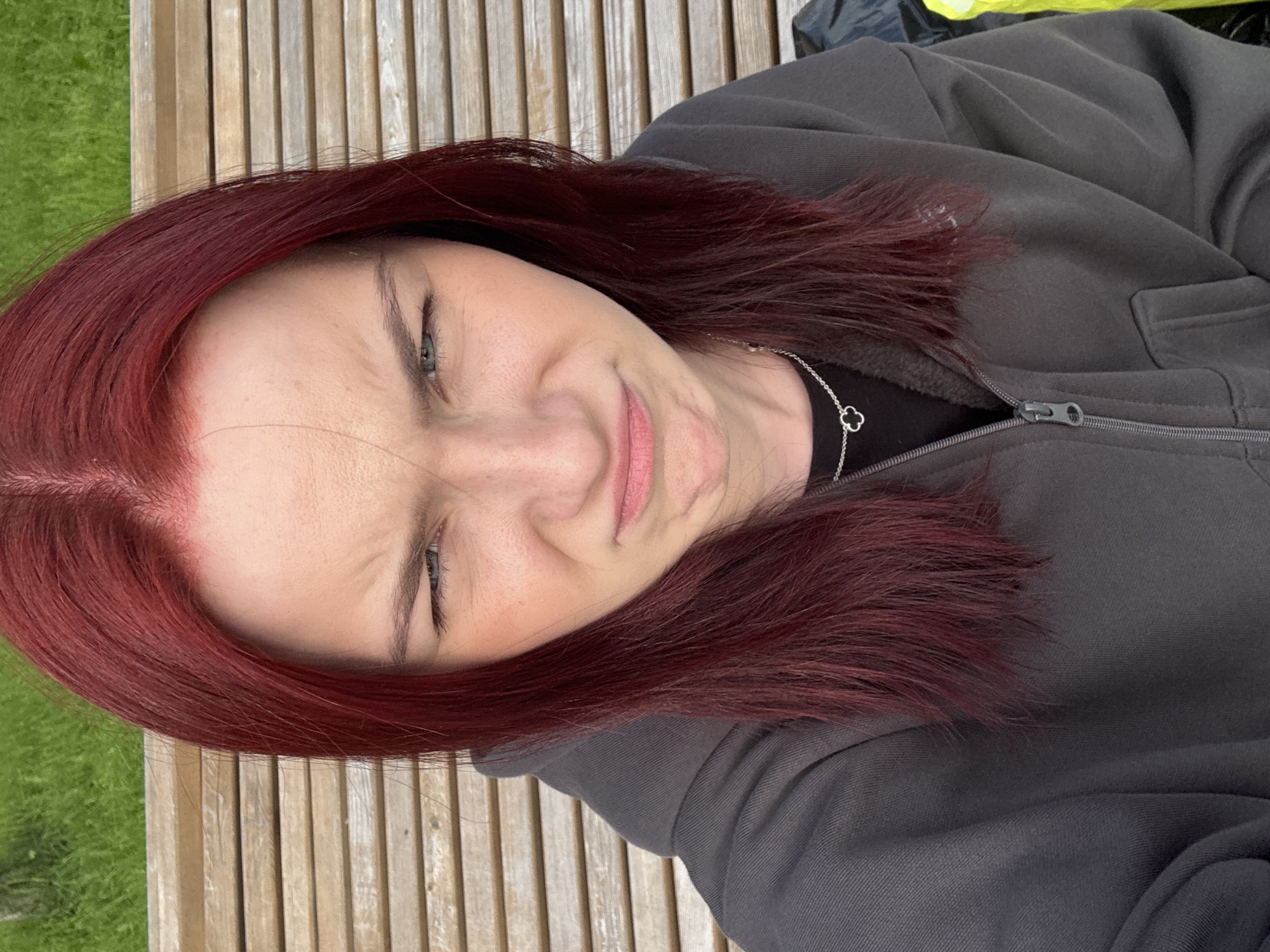 A young Caucasian female with dyed red hair and blue eyes is sitting on a wooden bench outdoors. She is wearing a dark grey zip-up hoodie over a black t-shirt. She has a pendant necklace with a clover-shaped charm. The background shows a grassy area and more wooden benches. The woman is looking directly at the camera with a disgruntled expression, her brow furrowed and her mouth pursed. The lighting suggests it is daytime. The location is unknown, and the country is Russia.