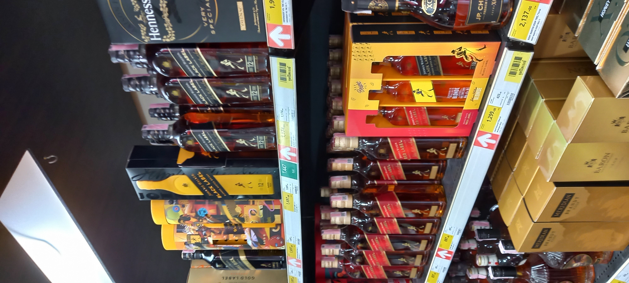 The image, rotated 90 degrees clockwise, captures a well-stocked aisle of alcoholic beverages in what appears to be a retail store, likely a supermarket or liquor store, in Moo 1, Thailand. The shelves are filled primarily with various brands and types of whiskey, brandy, and cognac.
On the left side of the frame, multiple bottles of Johnnie Walker Black Label whiskey are prominently displayed, stacked vertically. Price tags for these bottles show values of 1,174 and 1,697. Above these, a box for Hennessy Very Special Cognac is visible, with a partial price of 1,9__. Below the main stack of Black Label, there are special edition or gift-packaged Johnnie Walker Black Label products, including cylindrical packaging and boxes for Johnnie Walker Blonde, priced at 1,299.00, and another item at 433.
On the right side of the image, numerous bottles of Johnnie Walker Red Label are arranged on shelves, with a visible price of 697. Above them, boxed sets for Johnnie Walker Blonde and Johnnie Walker Red Label are stacked. Further up, a bottle of JP. Chenet French Brandy and a Remy Martin VSOP 70CL, priced at 2,137.00, are visible. Below the Red Label bottles, golden boxes for "Meridian Brandy" and "Baron Brandy" are neatly stacked, some with "HONEY" written on them.
The scene implies a retail environment focused on product display and sales. Many price tags feature an upward-pointing arrow and include Thai text, such as "จุดปิดเบย์" (Jut Pit Bay), which likely indicates a display point or special section, confirming the location in Thailand. The bottles and packaging are clean and well-organized, suggesting a commercial setting.