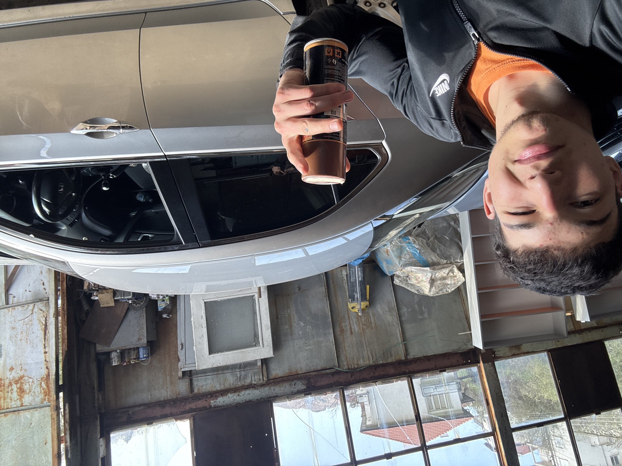 The image depicts a young man holding a cylindrical container with a brown bottom and dark-colored label. He's wearing a black Nike zip-up jacket over an orange shirt. He is positioned close to the camera, with a car and a garage visible in the background. The car is light-colored and parked inside the garage. The garage has rusty metal walls and broken windows looking out onto a residential area with tiled roofs. Based on the architectural style and general appearance of the environment, the location is consistent with being in Zlatograd, Bulgaria.
