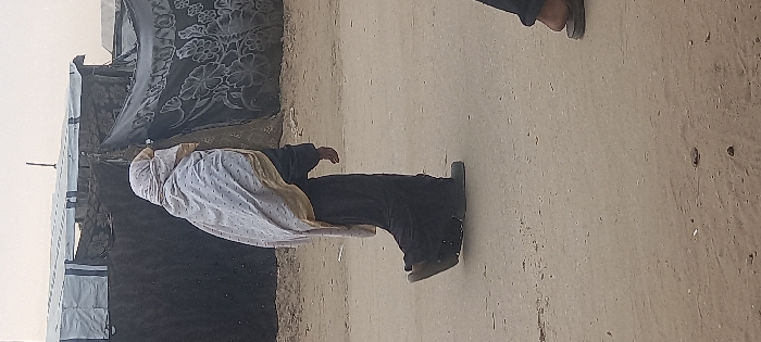 An individual, gender unidentifiable, walks from left to right on an unpaved dirt path in Khan Yunis, Palestinian Territory. The person wears a dark, long-sleeved garment and a light-colored head covering draped with a lighter patterned shawl. Their face is obscured. In the background, two improvised structures are visible: one on the left appears constructed from dark material with a light-colored roof section, and another on the right is covered with a dark fabric featuring a white floral pattern. A thin pole extends vertically from behind the structures. In the lower right foreground, a portion of a second person's foot, clad in a dark sandal, is visible. The sky is overcast.