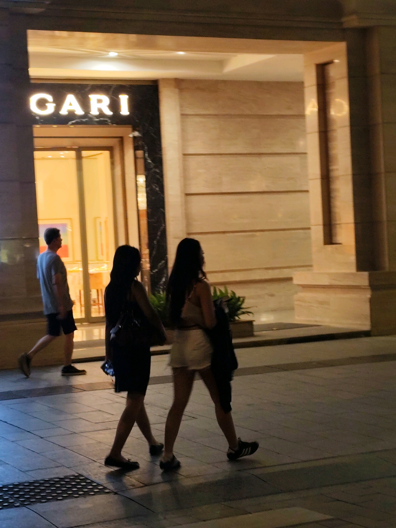 In Ho Chi Minh City, Vietnam, a man wearing a grey t-shirt and black shorts is walking past a store entrance. Two women, one in a black dress and the other in a white skirt and a dark top, are walking away from the viewer. The store has a large sign that reads "GARI". The scene appears to be at night, with artificial lighting illuminating the area.