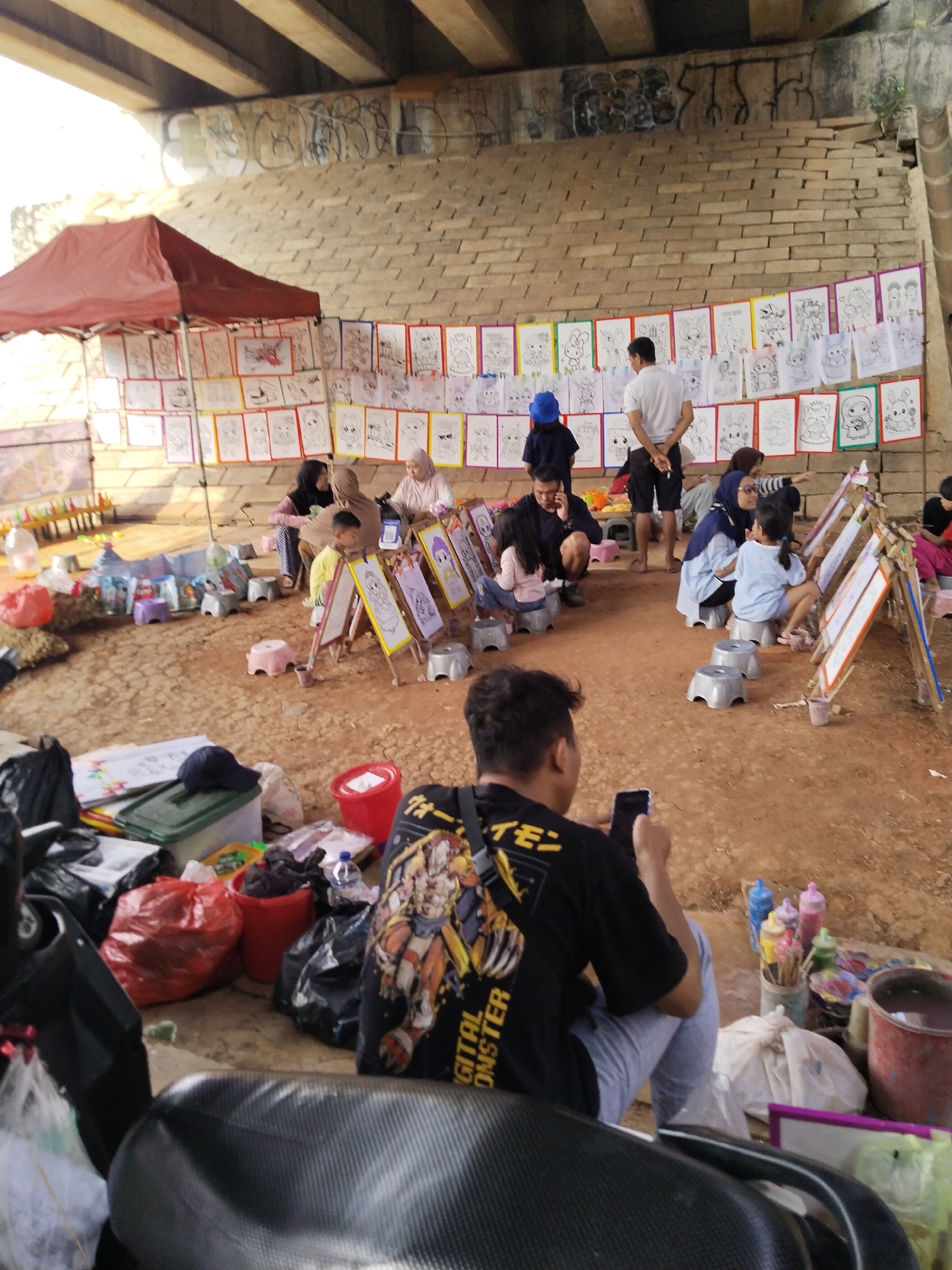Underneath a bridge with graffiti on the supporting beams, a community art activity is taking place. Rows of children's drawings, primarily in black and white with colored borders, are displayed on a brick wall. Several easels are set up on the sandy ground, where children and adults are engaged in drawing and painting. Various art supplies, including paint bottles, buckets, and bags of materials, are scattered around the area. In the foreground, a person is seated on a motorcycle, looking at their phone, with bags and other items near them. The scene suggests a vibrant outdoor event focused on fostering creativity among children.