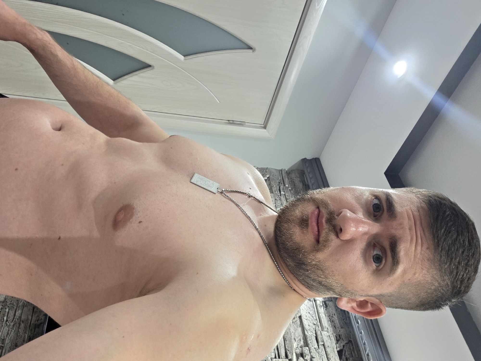 A rotated, close-up indoor portrait features an adult man, likely in Yagodovo, Bulgaria, looking directly at the camera with a neutral and somewhat pensive expression. The man has short brown hair, a well-groomed beard, and light-colored eyes. He is shirtless, with his bare torso and right arm visible, suggesting an intimate or casual setting. He wears a silver-colored chain necklace with a rectangular dog-tag style pendant; faint, indecipherable text or a design is etched onto its surface.
The background elements suggest a modern interior. On the left, a light-colored wooden door with a frosted glass panel is visible. To the right, a wall covered in textured material, possibly stone or brick wallpaper, adds depth. Above, a bright recessed light fixture on the white ceiling casts a strong beam, indicating artificial lighting within the room. A dark architectural detail, possibly a beam or frame, is also present in the upper right portion of the frame. The image appears to be a self-portrait or a photograph taken from a low angle.