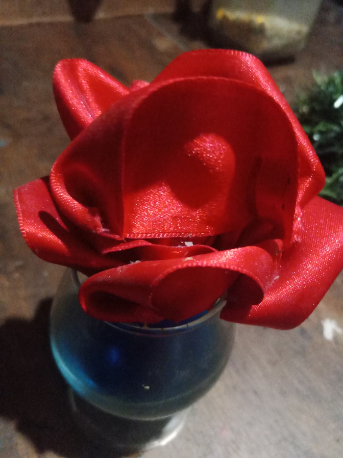 A red, ribbon-like rose fills the frame, its satin texture reflecting light. The rose is set in a small, rounded vase filled with blue liquid. The scene is illuminated by a soft, warm light. To the upper right is a bit of green plant or brush. The vase and the rose sit on a wood-like surface.