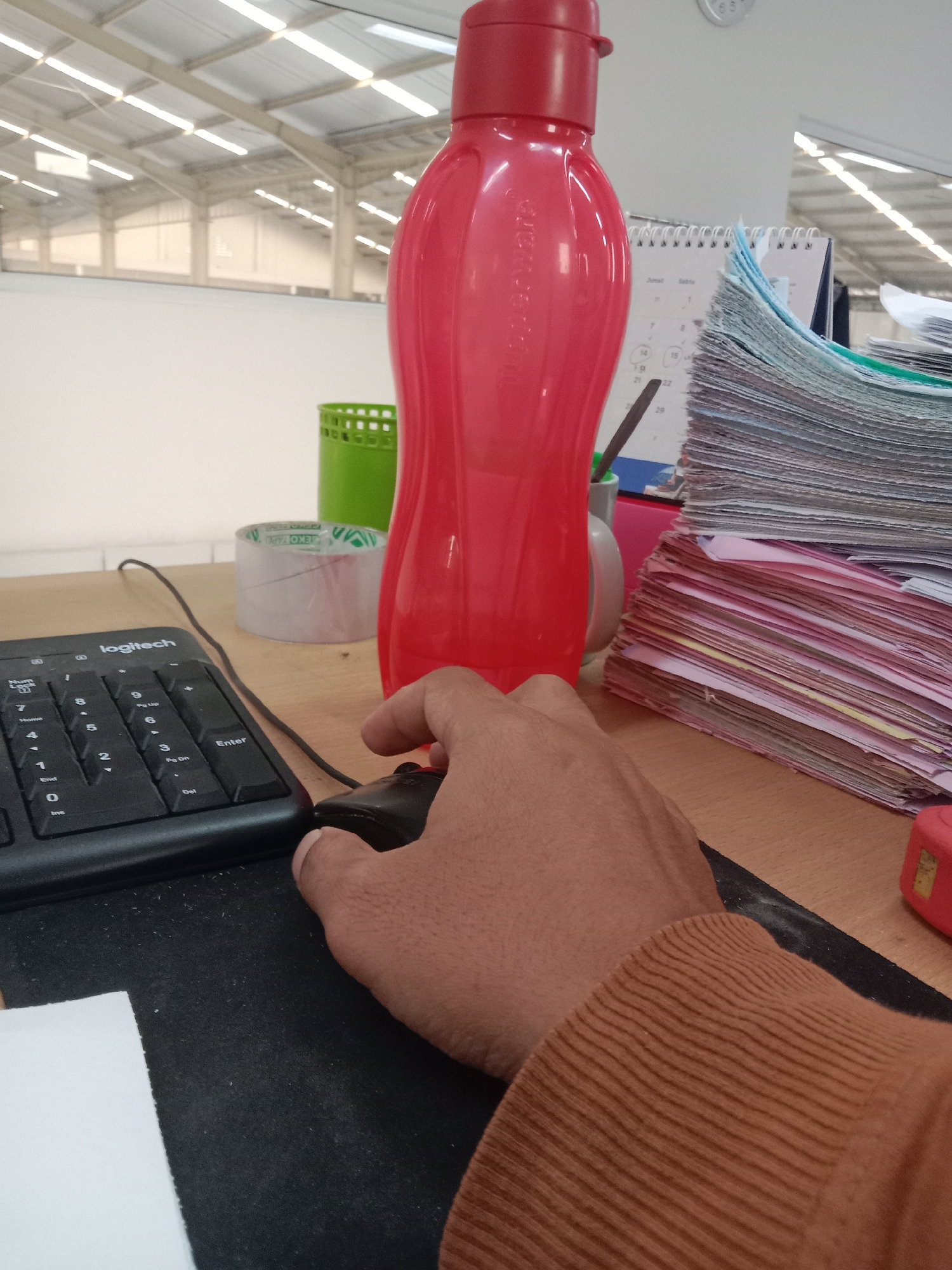 The image depicts a desk in what appears to be an office environment within a large industrial building, possibly located in Semarang, Indonesia. The background shows a high ceiling with rows of fluorescent lights and structural beams, characteristic of a warehouse or factory setting.
On the desk, a person's hand is seen operating a black computer mouse. In front of the mouse is a black Logitech keyboard. To the right of the keyboard, a stack of papers and documents are piled high, with some appearing to be calendar pages. A red Tupperware-style water bottle stands prominently on the desk, and a roll of clear tape is visible to its left. A green cylindrical holder, possibly for pens or other office supplies, is also present.
The overall scene suggests a workspace where work is in progress, with the primary focus on the interaction between the person's hand, the computer mouse, and the keyboard. The lighting suggests an indoor environment, possibly during daylight hours as indicated by the illumination from the overhead lights. There is no direct evidence to pinpoint the exact location within Semarang, but the architectural style of the building suggests an industrial or commercial area.