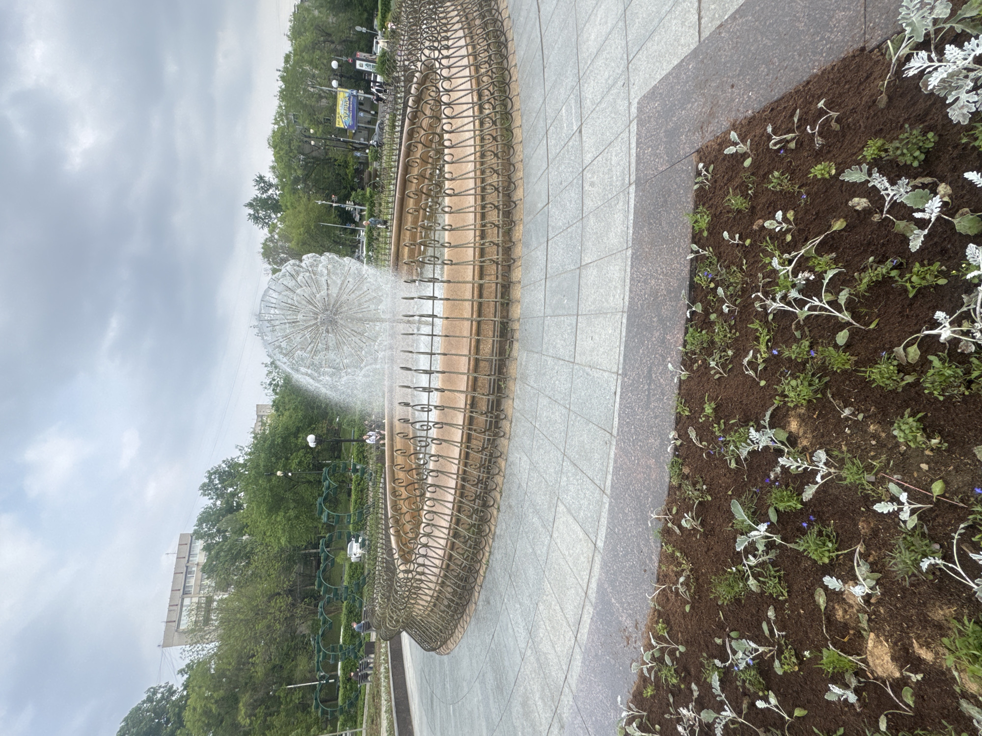 The image depicts a fountain in Khabarovsk, Russia. The fountain features a spherical water spray in the center, surrounded by an ornate, circular metal fence. The area around the fountain is paved with gray tiles, and there is a flowerbed with silver-leaved plants adjacent to the pavement. Trees and buildings are visible in the background under a cloudy sky.
