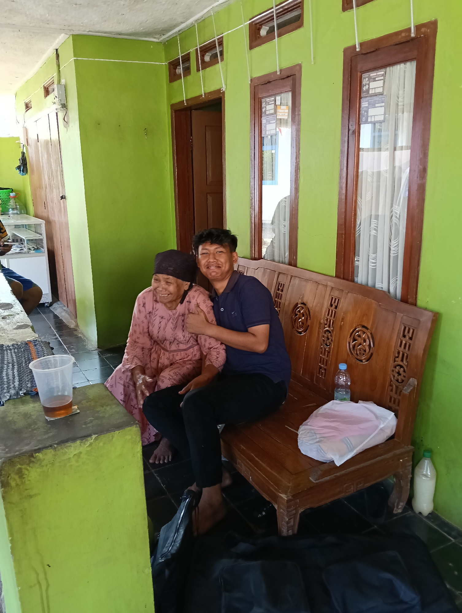 The image shows a young man with dark hair and a dark blue polo shirt, and an elderly woman with a dark headscarf and a pink dress, sitting on a wooden bench. The man has his arm around the woman's shoulders and is giving a thumbs-up. The woman is smiling and looking to her left. They are sitting in front of a bright green wall with two wooden-framed windows. The bench is ornately carved. To the left of the bench, there is a low, bright green concrete counter with a plastic cup of amber liquid and a woven mat. In the background, there is a wooden door and another window. On the floor, there are black bags. A plastic bottle of water and a plastic bottle with a light-colored liquid are visible near the bench. The scene appears to be an outdoor or semi-outdoor area of a residence in Tasikmalaya, Indonesia, given the architectural style, the presence of tropical plants (implied by the bright green walls, common in Indonesian homes), and the general ambiance suggesting a warm climate.
