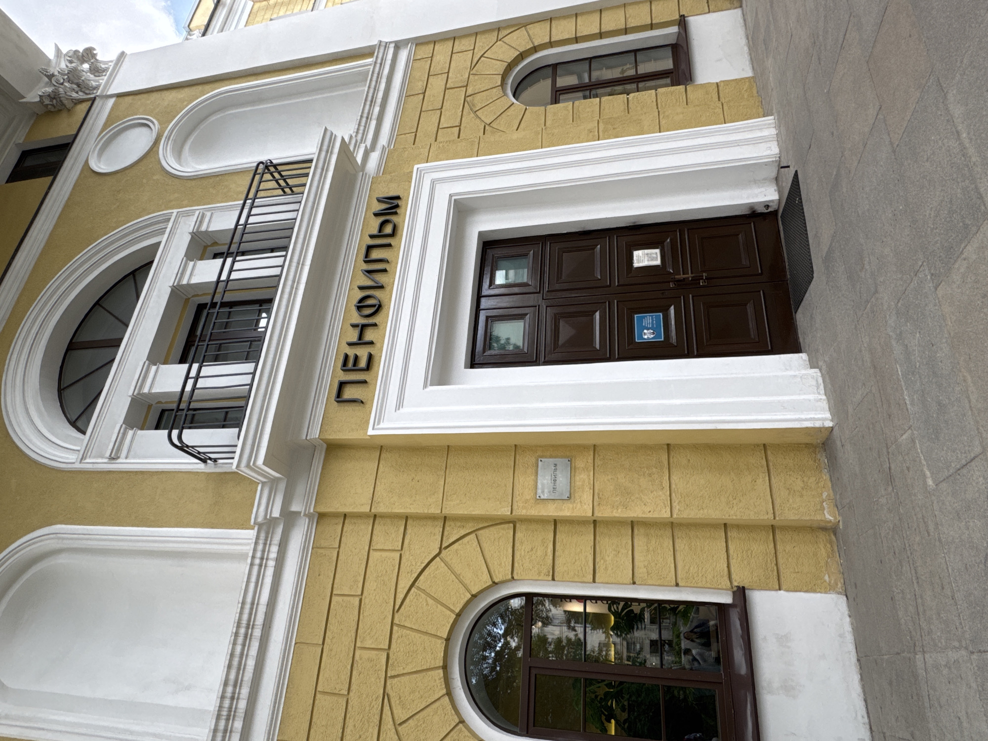 The image shows the exterior of a building with a yellow facade and white architectural details. Above a dark brown double door entrance, large black Cyrillic letters spell out "ЛЕНФИЛЬМ" (LENFILM). To the left of the entrance is a round-arched window with dark framing, reflecting trees and buildings. To the right of the entrance, there is a smaller rectangular window, also with dark framing. Above the entrance, a balcony with a black metal railing is visible. A rectangular metal plaque with Cyrillic text is affixed to the wall to the left of the entrance. The building's architecture suggests it is an older, possibly historical, structure. The ground in front of the building is paved with grey stone tiles. The scene appears to be a street in a city, likely Saint Petersburg, Russia, given the prominent signage of "Lenfilm," a historic film studio.