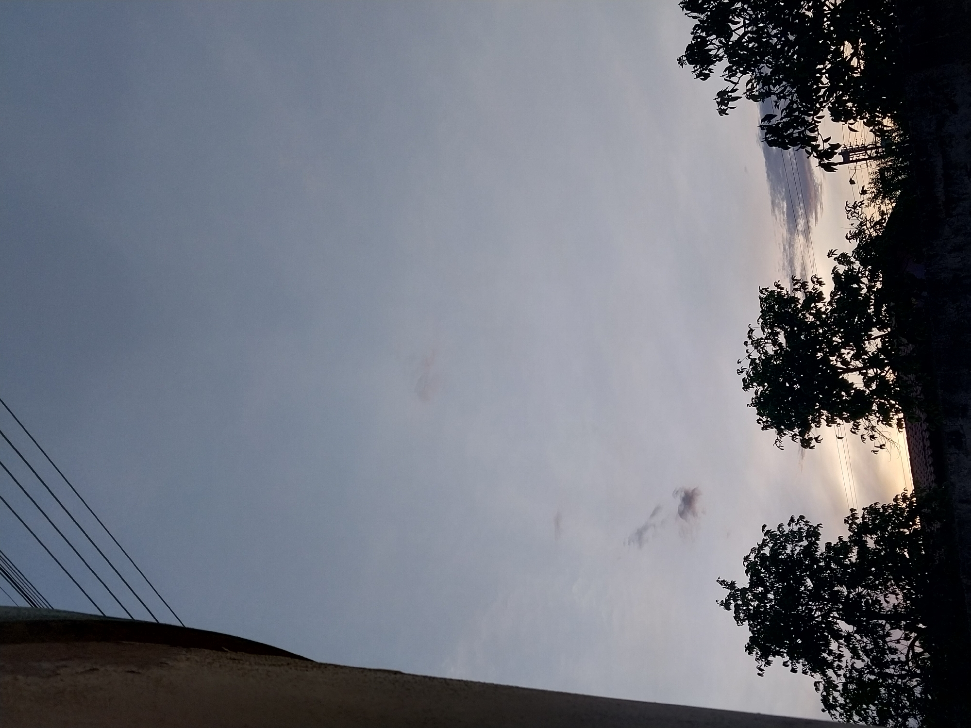 The image depicts a sky at dusk in Tinaquillo, Venezuela. The sky is predominantly a light grey color, with hints of yellow and orange near the horizon, suggesting a sunset or sunrise. The silhouettes of trees are visible on the right side of the frame, and overhead power lines extend from the bottom left corner. The top of a building is visible in the bottom left.